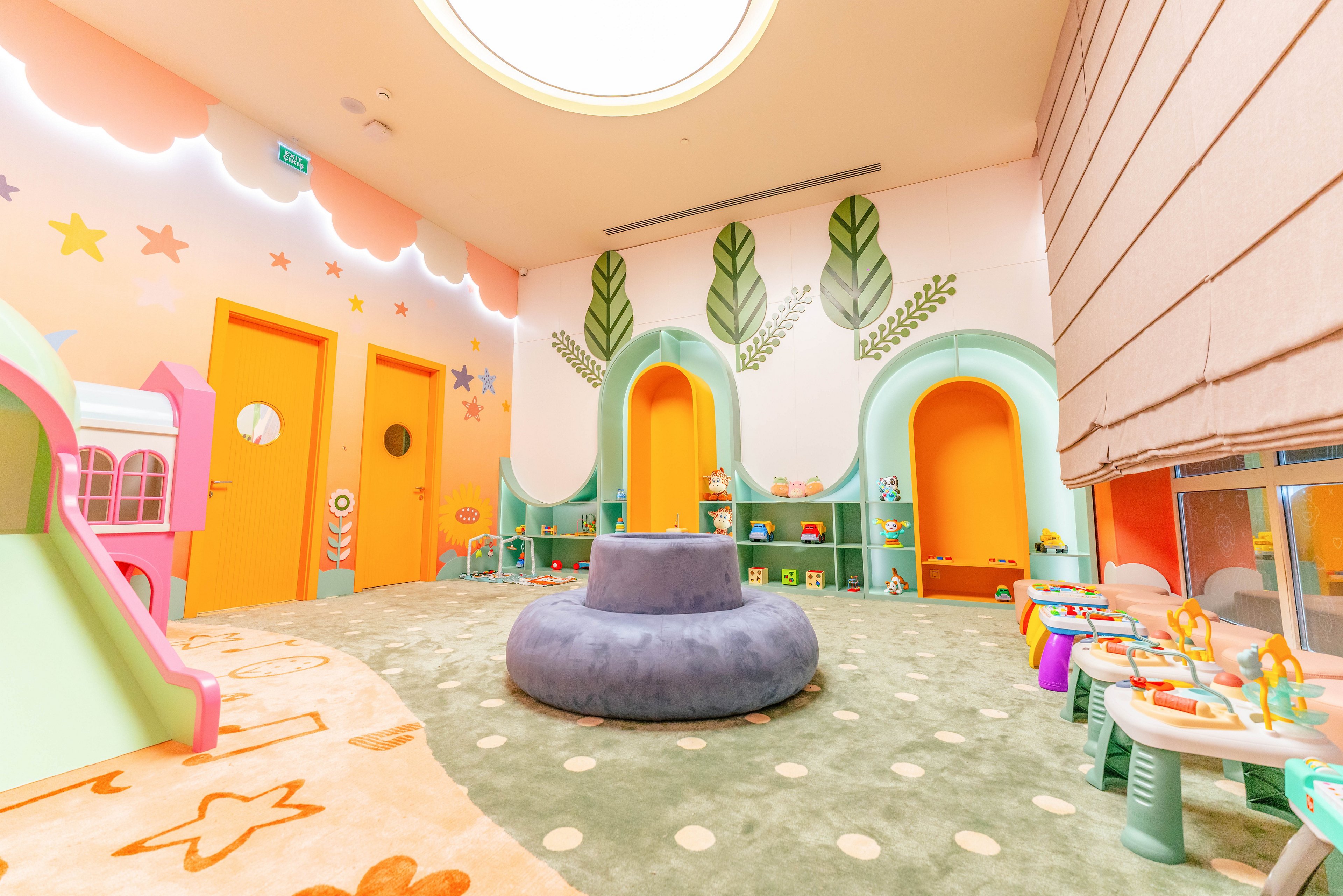 A vibrant children's playroom features pastel walls decorated with stars, leaves, and whimsical patterns. The space includes multiple yellow doors, a plush circular seating area, and various play tables with toys. The room is well-lit with a large ceiling light and has a cozy, inviting atmosphere. No visible text or numbers are present in the image.