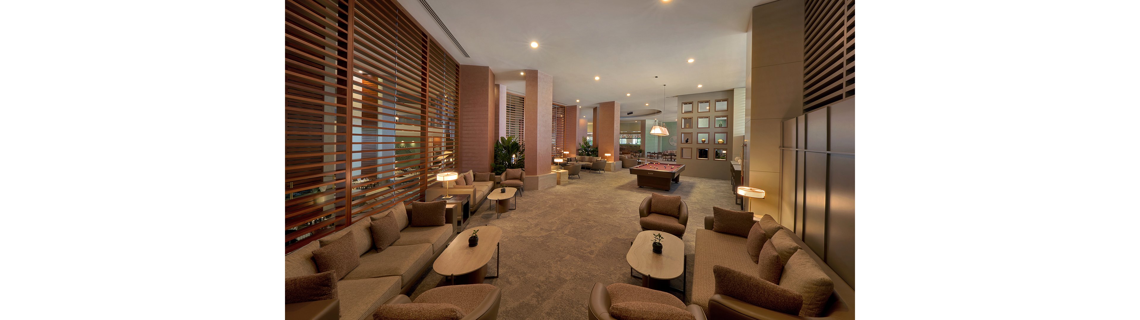 A large, modern lounge features multiple seating areas with cushioned sofas and armchairs arranged around low coffee tables. Warm lighting and wooden slat partitions create an inviting atmosphere in the open-plan space. In the background, a billiards table and wall art add recreational and decorative elements. The overall design emphasizes comfort, relaxation, and social interaction in a refined interior setting.