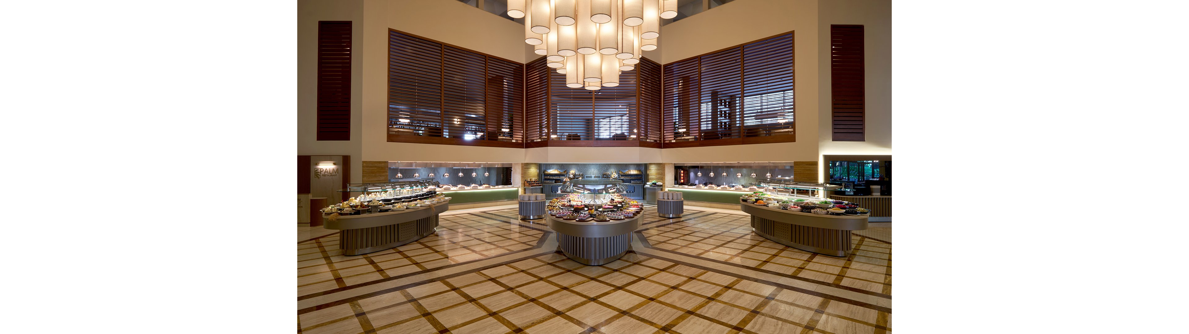 Wide view of an elegant buffet restaurant with multiple circular food stations arranged in the center of a large tiled floor. The room features high ceilings, warm lighting, and modern pendant lamps hanging above the buffet islands. Various dishes and desserts are neatly presented along the central stations and back counters. No people are present, emphasizing the architecture, symmetry, and abundance of food options.