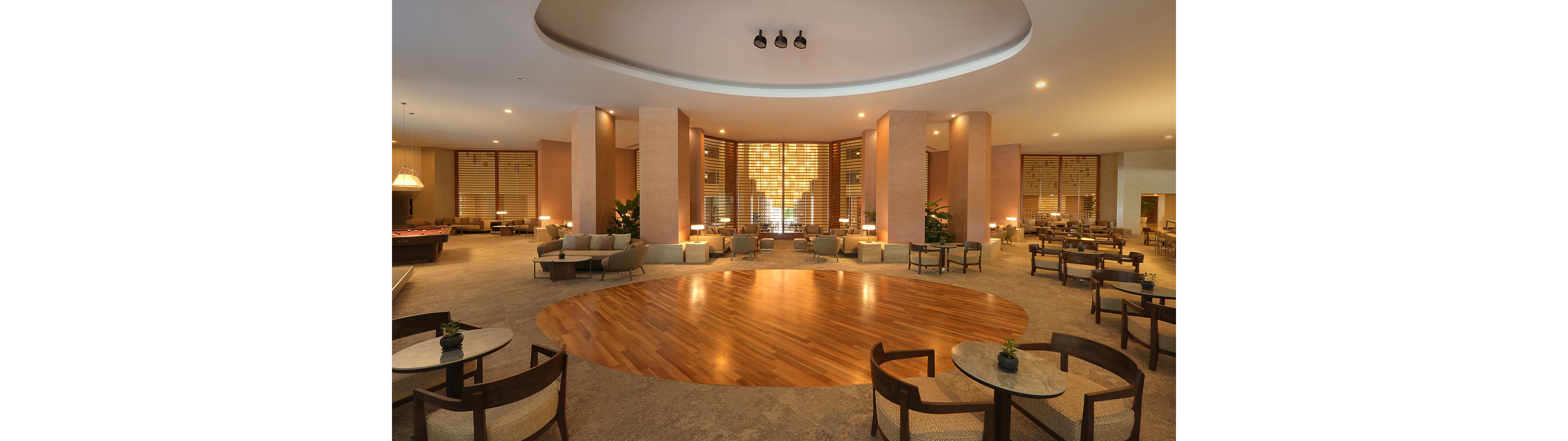 Large open lobby lounge with modern seating surrounds a circular wooden floor feature. Soft warm lighting highlights tall columns and decorative wall panels in the background. Multiple tables and armchairs are arranged for socializing or relaxation, with small plants adding a touch of greenery. The overall design feels upscale, calm, and inviting, suitable for a hotel or resort setting.