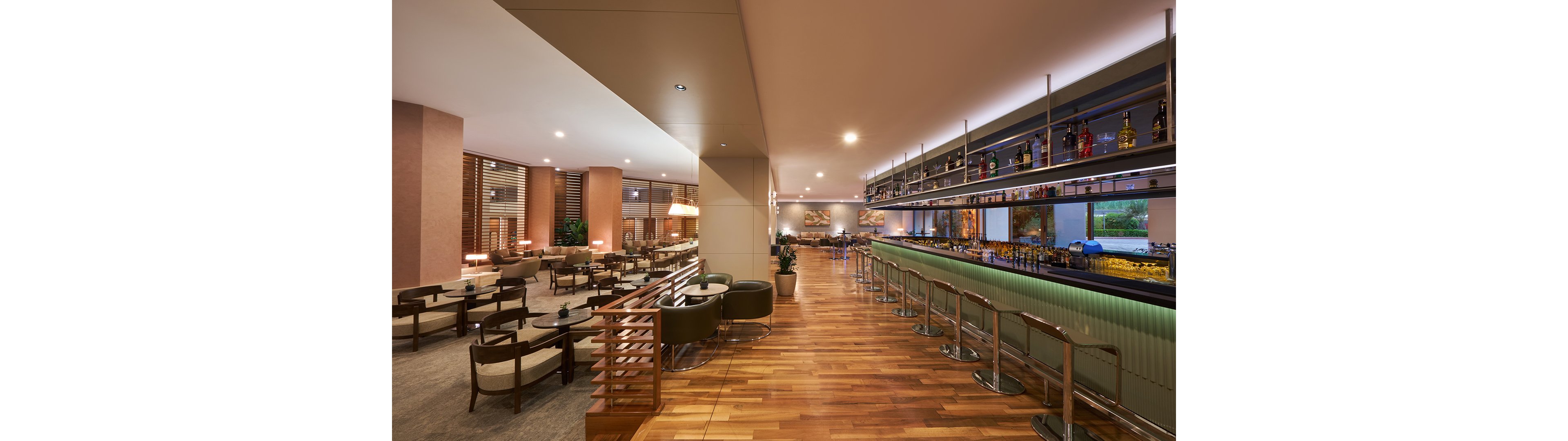 Spacious contemporary bar lounge with a long counter and high stools stretches across the right side of the image. On the left, comfortable armchairs and low tables create a relaxed seating area under warm ambient lighting. Bottles and glassware are neatly arranged on shelves above the bar, with large windows providing natural light and an outside view. The overall atmosphere is modern, inviting, and designed for socializing or casual meetings.
