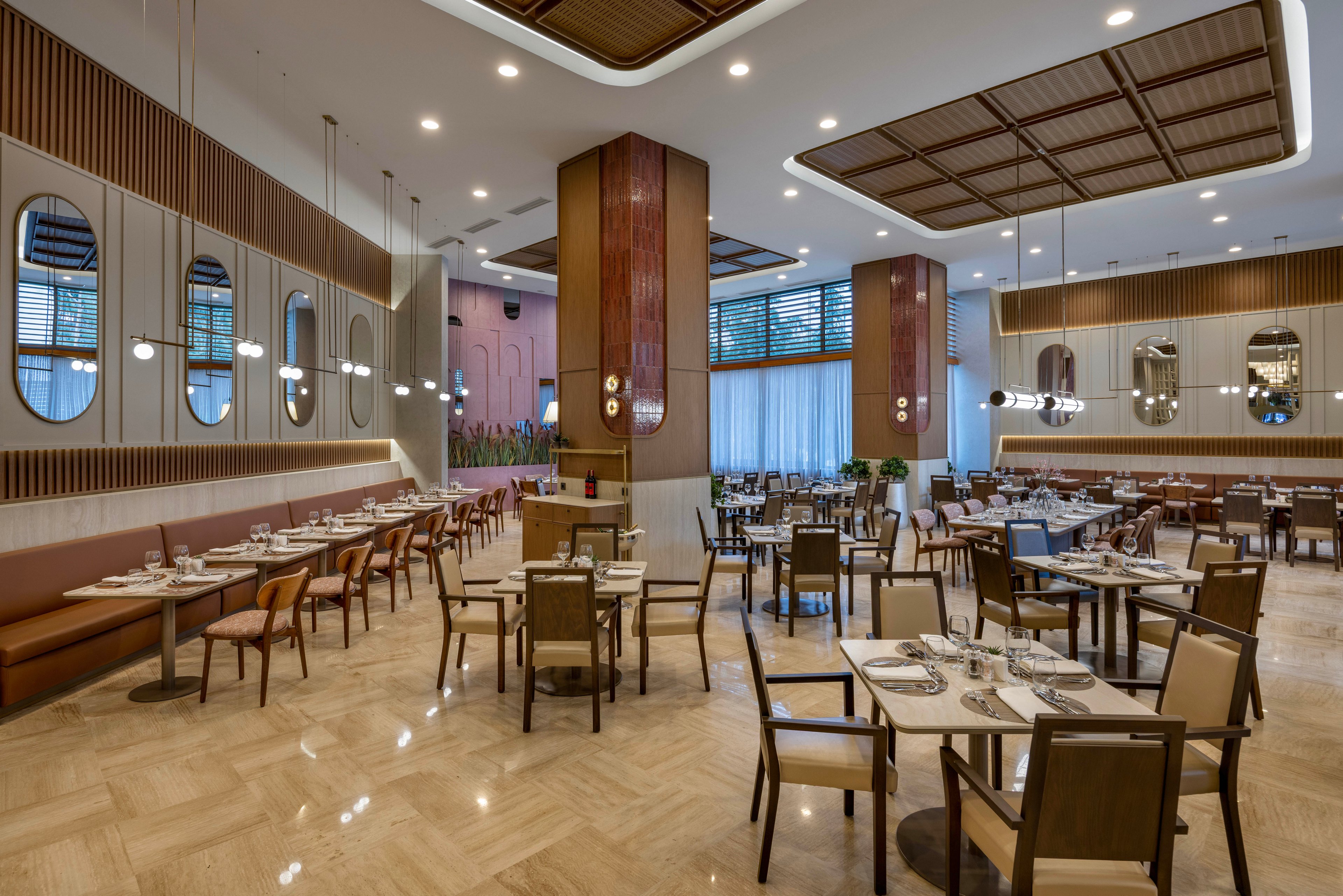 Spacious restaurant dining area featuring neatly arranged tables and chairs, set for service. The setting includes contemporary decor with large mirrors, pendant lighting, and wood accents. The atmosphere is bright and inviting, with neutral tones and polished flooring. No people are present, highlighting the clean and organized environment.