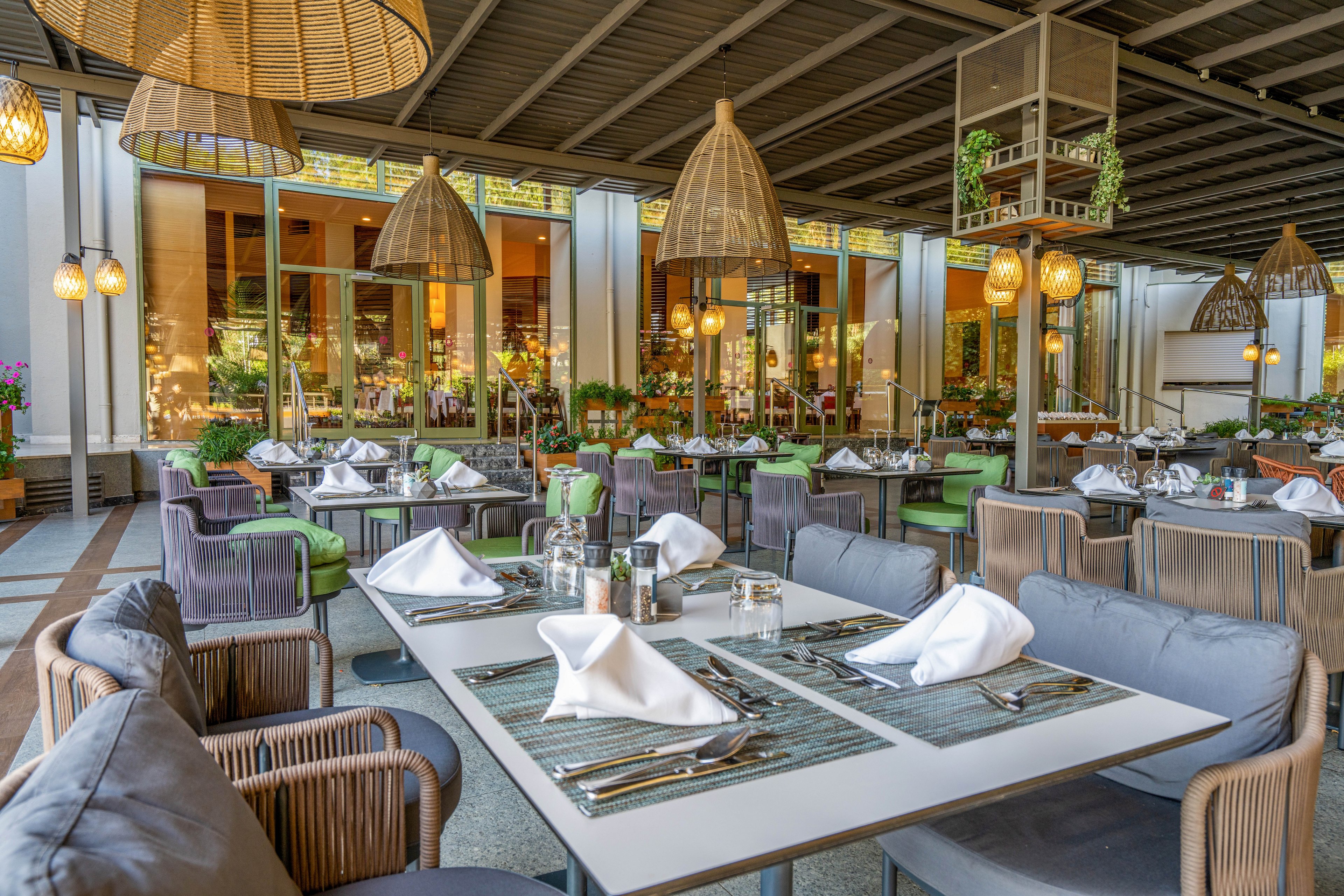 A stylish outdoor dining area features neatly arranged tables with white napkins, glassware, and cutlery. The setting is under a covered patio with woven pendant lights and comfortable cushioned chairs. Lush greenery and potted plants add a fresh ambiance, while large windows reflect the inviting atmosphere. The overall mood is relaxed and upscale, ideal for social gatherings or fine dining.