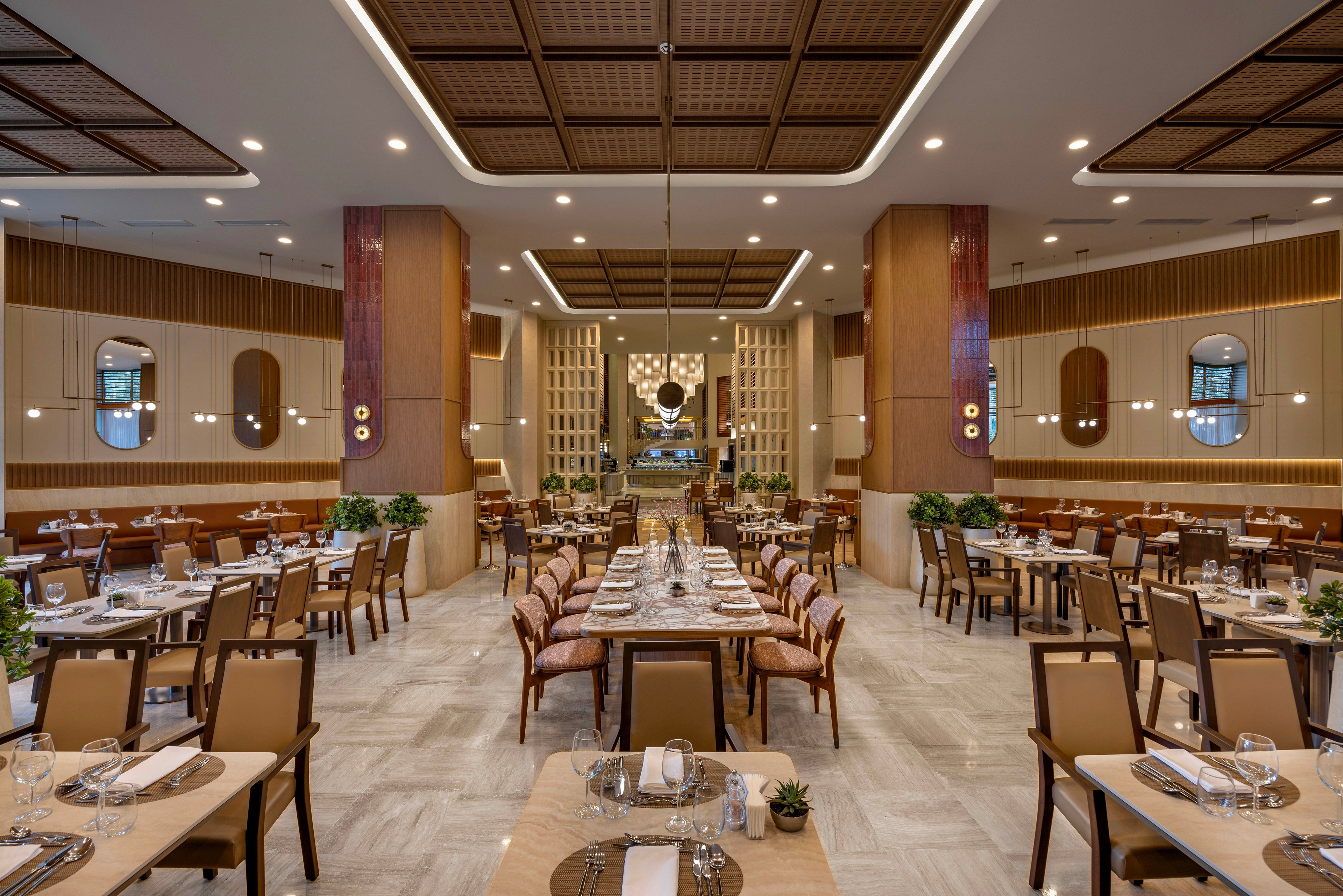 The image showcases a spacious, contemporary restaurant dining area with neatly arranged tables and chairs. The setting features warm lighting, wooden accents, and decorative plants, creating a welcoming and sophisticated atmosphere. Place settings with glassware and cutlery are visible, emphasizing readiness for guests. The overall mood is refined and inviting, with neutral and earthy tones dominating the palette. No visible text or numeric values are present in the image.