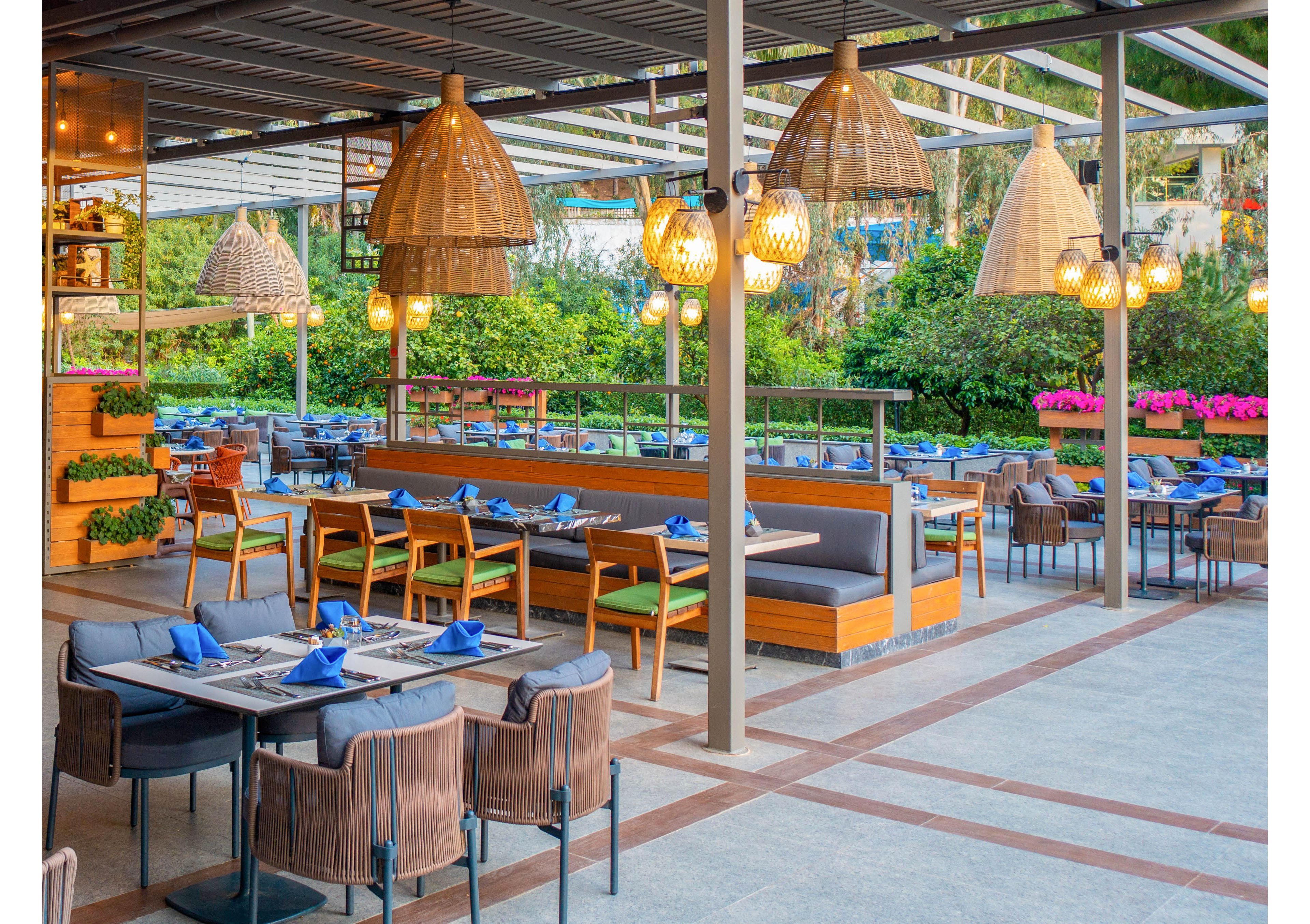 A contemporary outdoor restaurant features neatly arranged tables and cushioned booths, each set with blue napkins. The setting is bright and inviting, with large wicker pendant lights and lush greenery surrounding the dining area. The open-air design and colorful flowers create a relaxed, welcoming atmosphere. No people are present, emphasizing the tranquil and ready-to-serve environment.