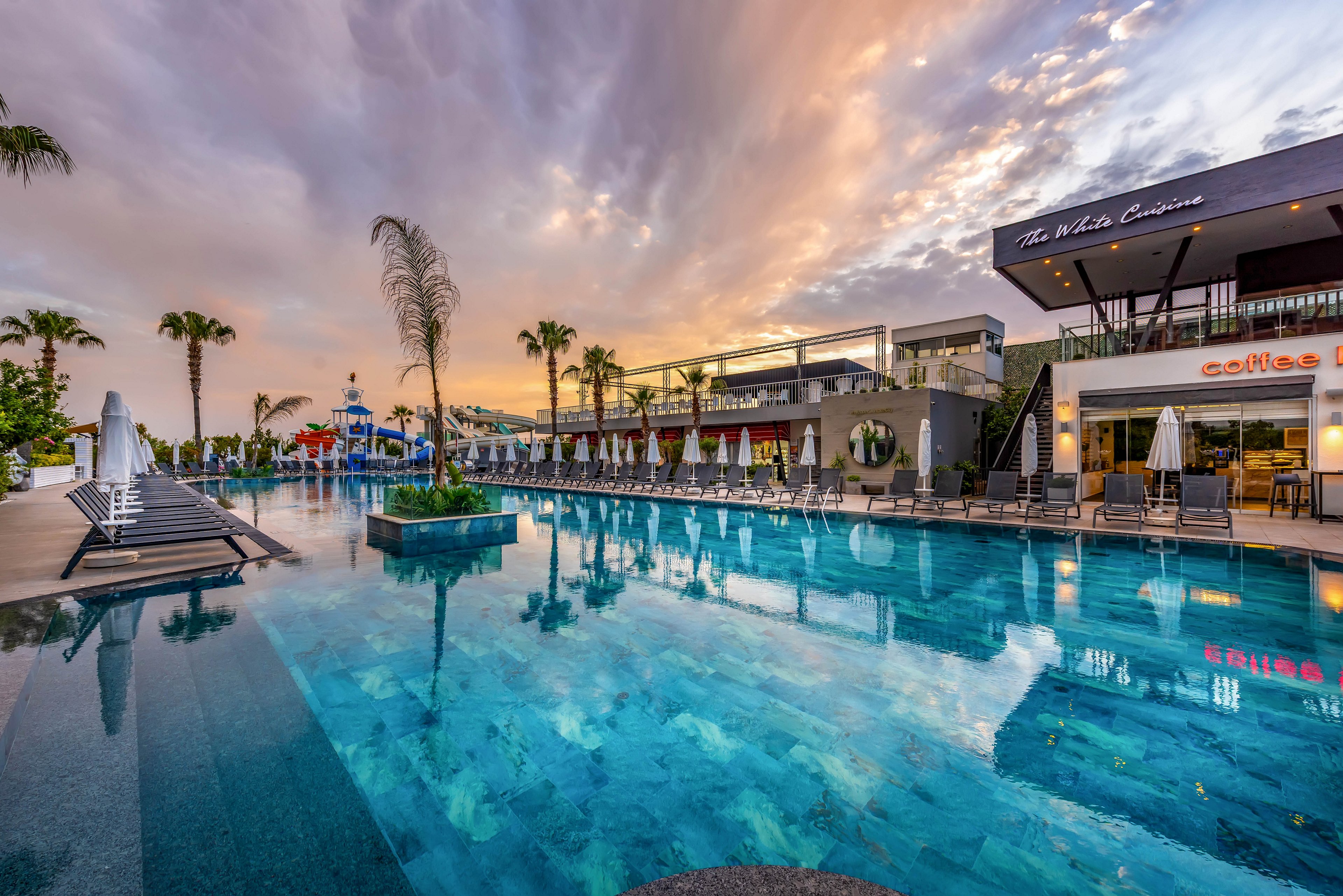 A large, tranquil swimming pool reflects the colorful sunset sky at a modern resort. White sun loungers and umbrellas line the poolside, with palm trees adding a tropical atmosphere. Contemporary buildings and outdoor dining areas frame the water, creating a stylish vacation setting. The calm water and warm evening light evoke a relaxed, upscale holiday mood.