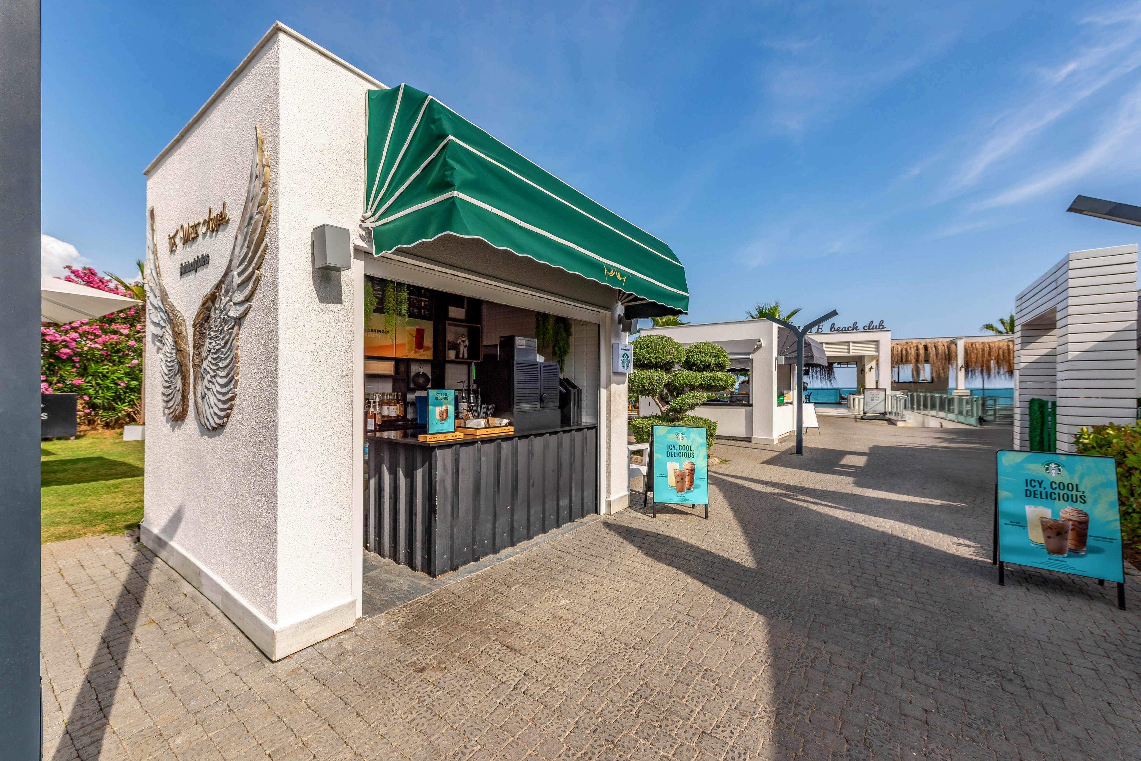 A small white coffee kiosk with a green awning stands along a sunny outdoor pathway. Promotional signs with iced coffee images are placed in front of the counter. Modern cabanas, manicured greenery, and a pool area are visible in the background, suggesting a resort or beach club setting. The scene is bright and relaxed, with clear blue skies and warm daylight.