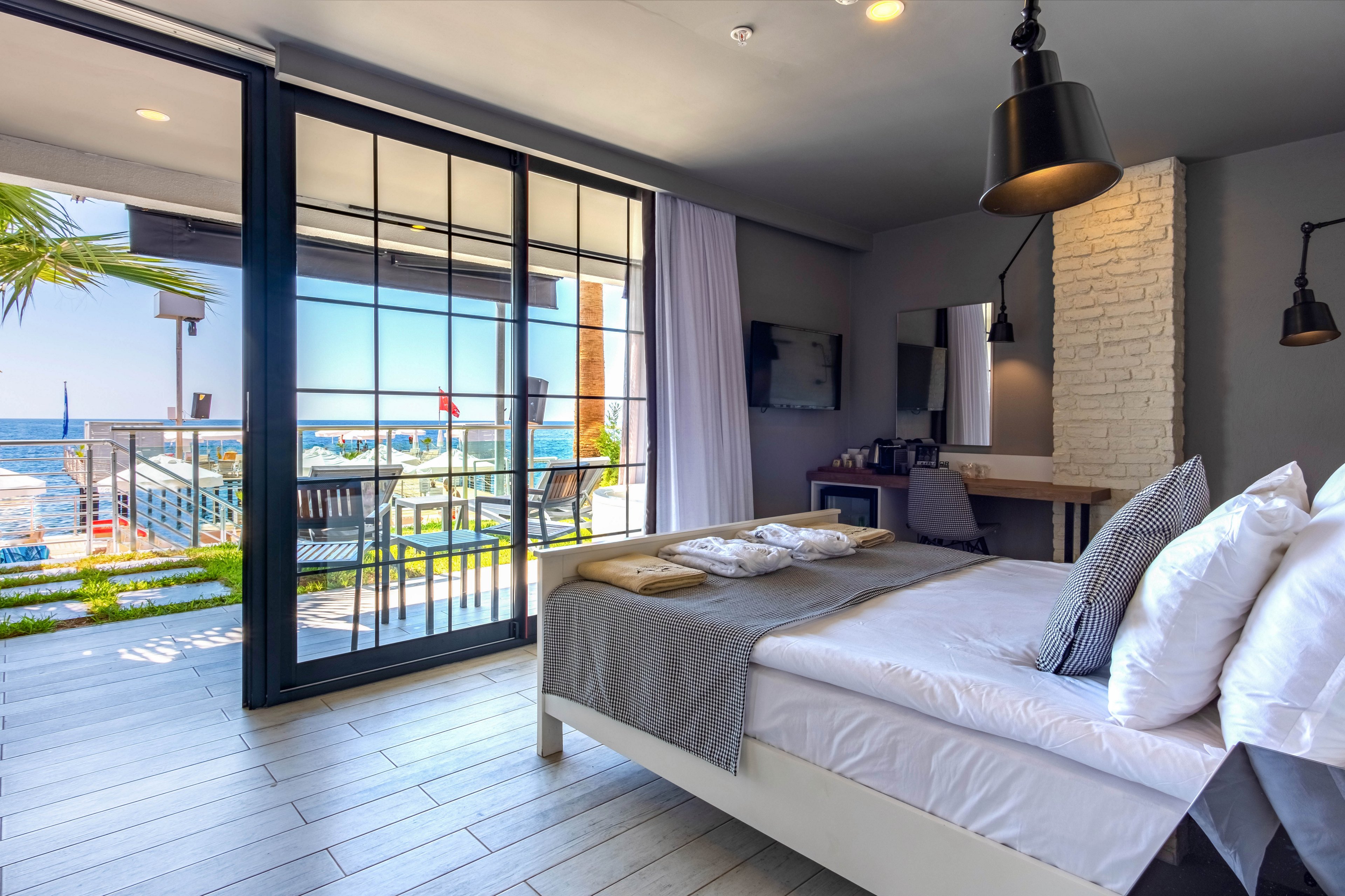Contemporary hotel room featuring a neatly made double bed faces large floor-to-ceiling glass doors. The room opens onto a balcony with chairs and a table overlooking the sea and a distant sailboat. Interior details include pendant lights, a white brick accent wall, a desk with a mirror, and a wall-mounted television. Natural light and neutral tones create a bright, relaxing coastal atmosphere.