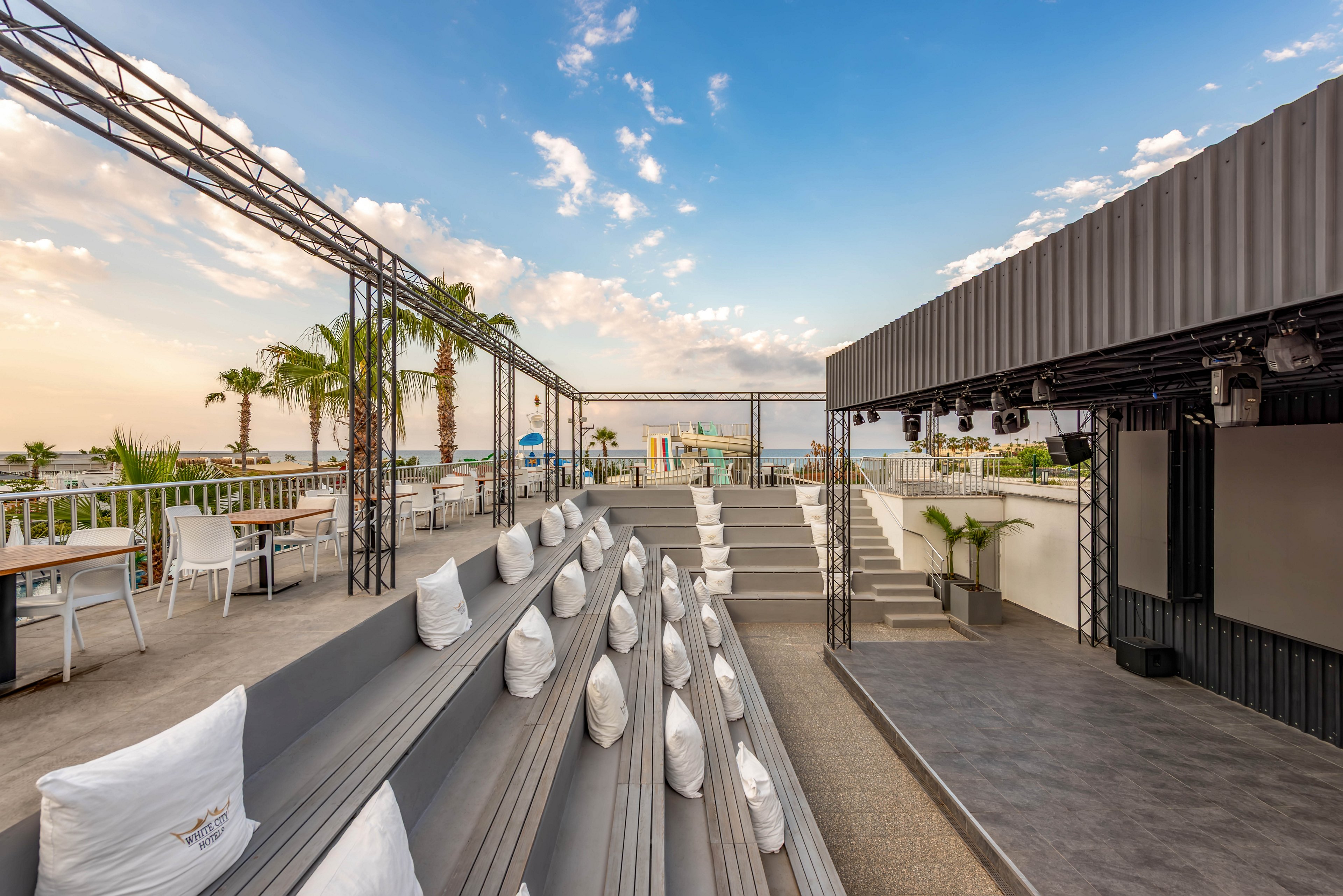 A contemporary rooftop terrace features gray tiered seating lined with white cushions. The space includes an open-air stage or event area framed by metal structures and a covered section on the right. Outdoor dining tables and chairs are arranged along the railing, overlooking a city or coastal skyline with palm trees. Soft evening light and scattered clouds create a relaxed, inviting atmosphere suitable for events or gatherings.