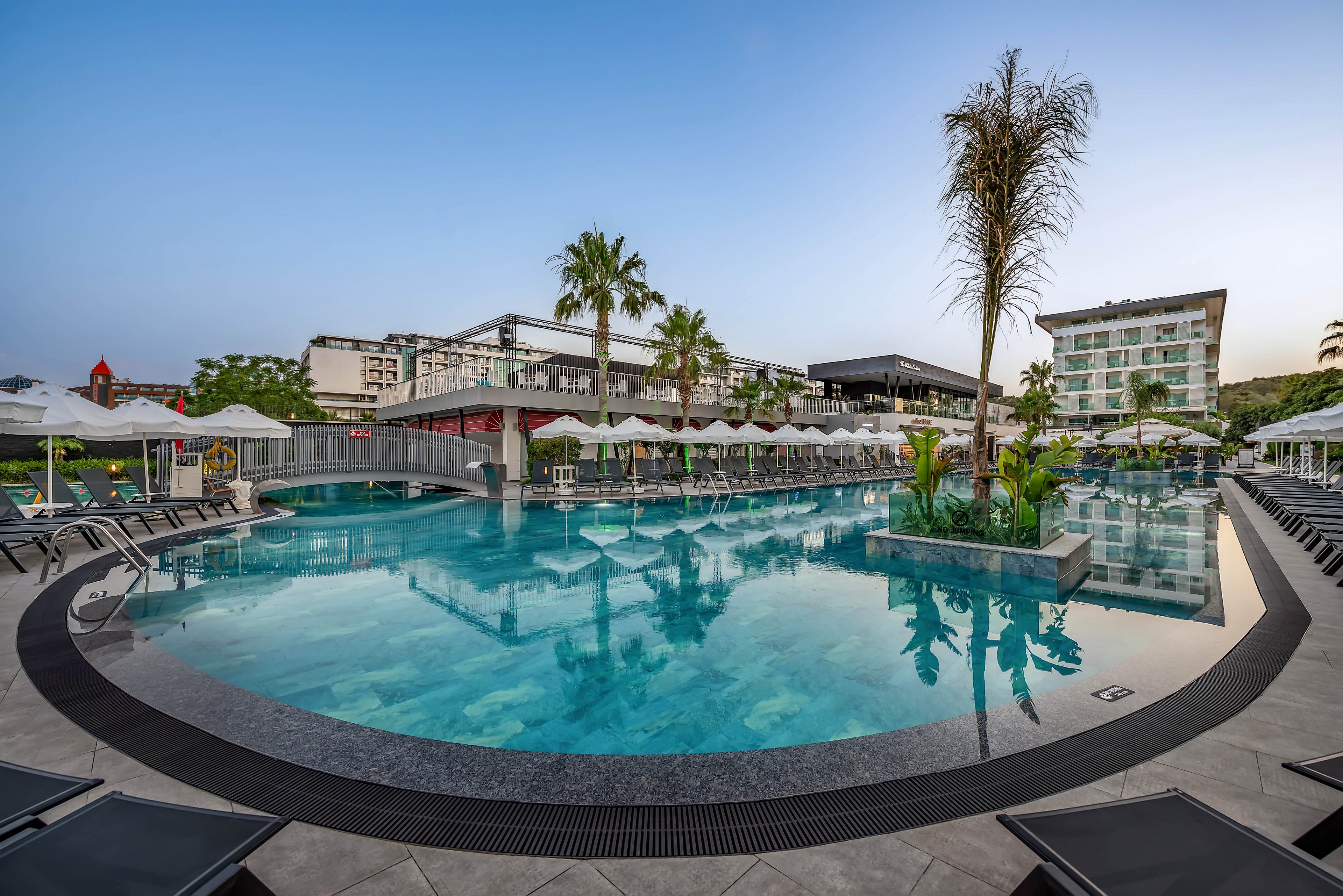 A large, curved swimming pool dominates the foreground of a modern resort complex. Sun loungers and white umbrellas line the poolside, creating a relaxed vacation atmosphere. A small bridge spans part of the pool, while palm trees and landscaped greenery add a tropical touch. Contemporary multi-story hotel buildings frame the scene under a clear early-morning sky.