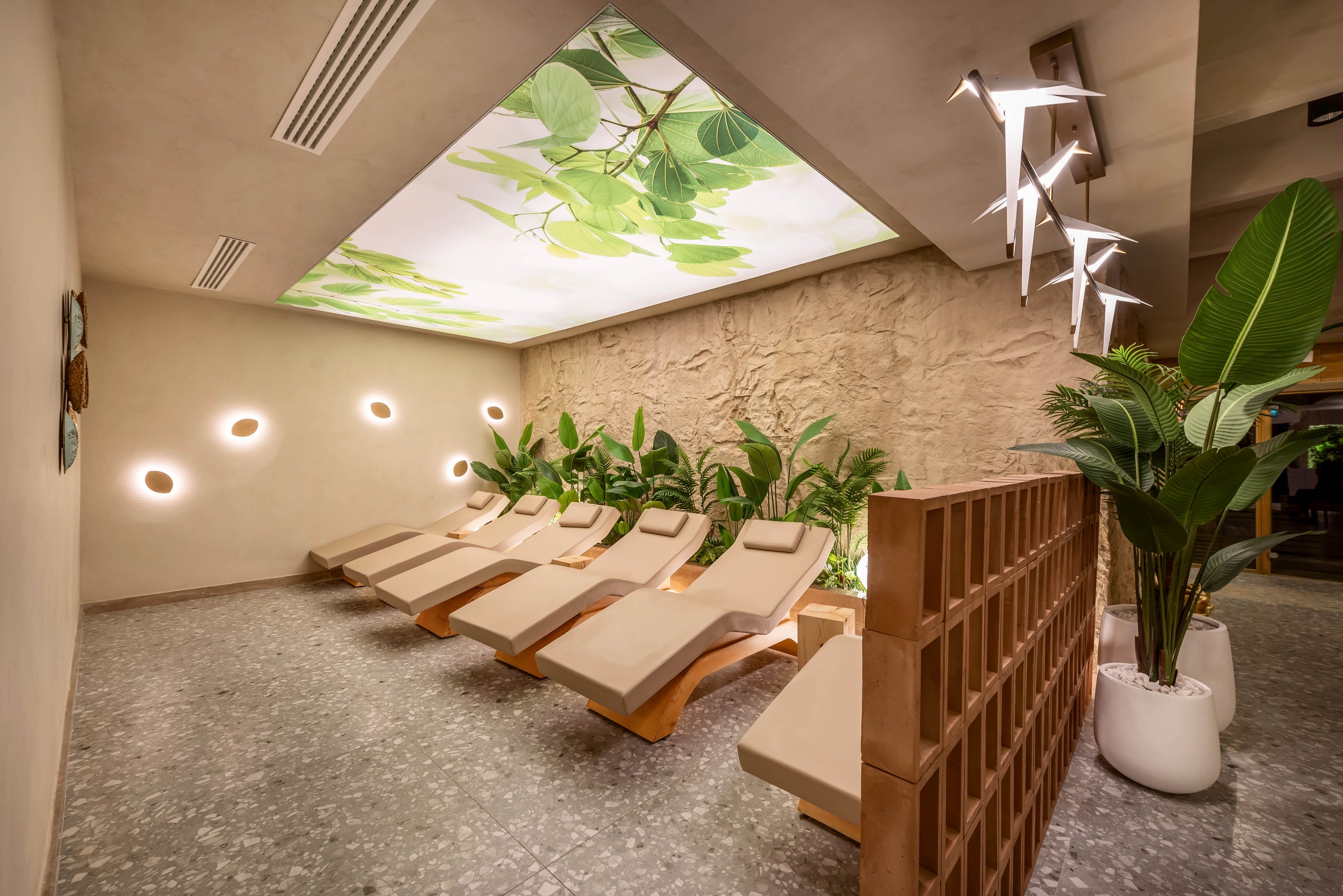 A contemporary spa relaxation lounge features a row of cushioned reclining loungers arranged along a textured wall. Soft circular wall lights and a large ceiling panel with green leaf artwork create a calming, nature-inspired atmosphere. Potted tropical plants and a decorative partition add warmth and privacy to the minimalist interior. The overall setting feels serene and inviting, ideal for rest and wellness.