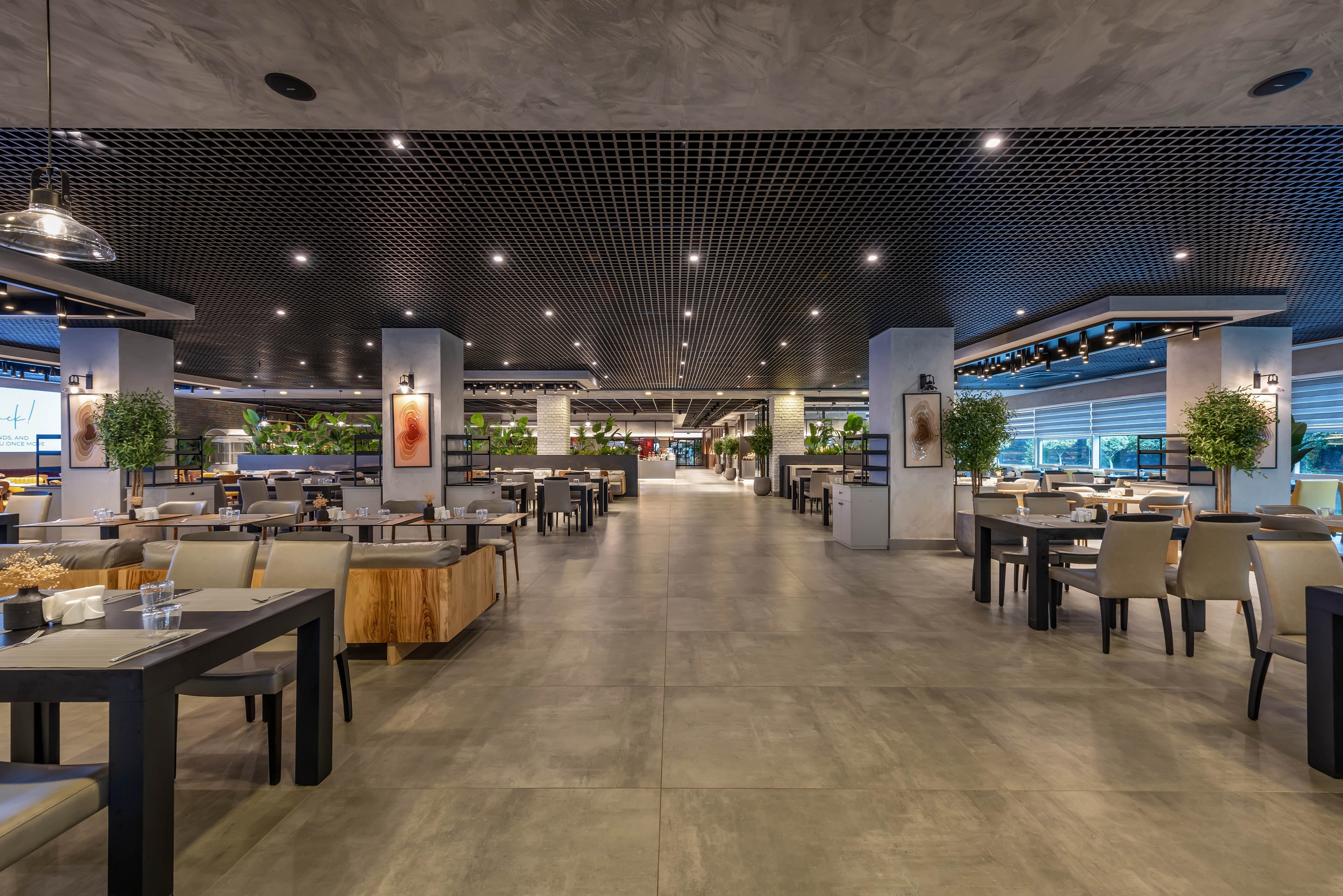A wide, modern restaurant dining hall features neatly arranged tables and chairs in a symmetrical layout. The space is brightly lit with a dark grid ceiling and large floor tiles creating a clean, contemporary look. Decorative plants and framed artwork add warmth and color along the central aisle. The restaurant appears empty, emphasizing the open floor plan and ample seating.
