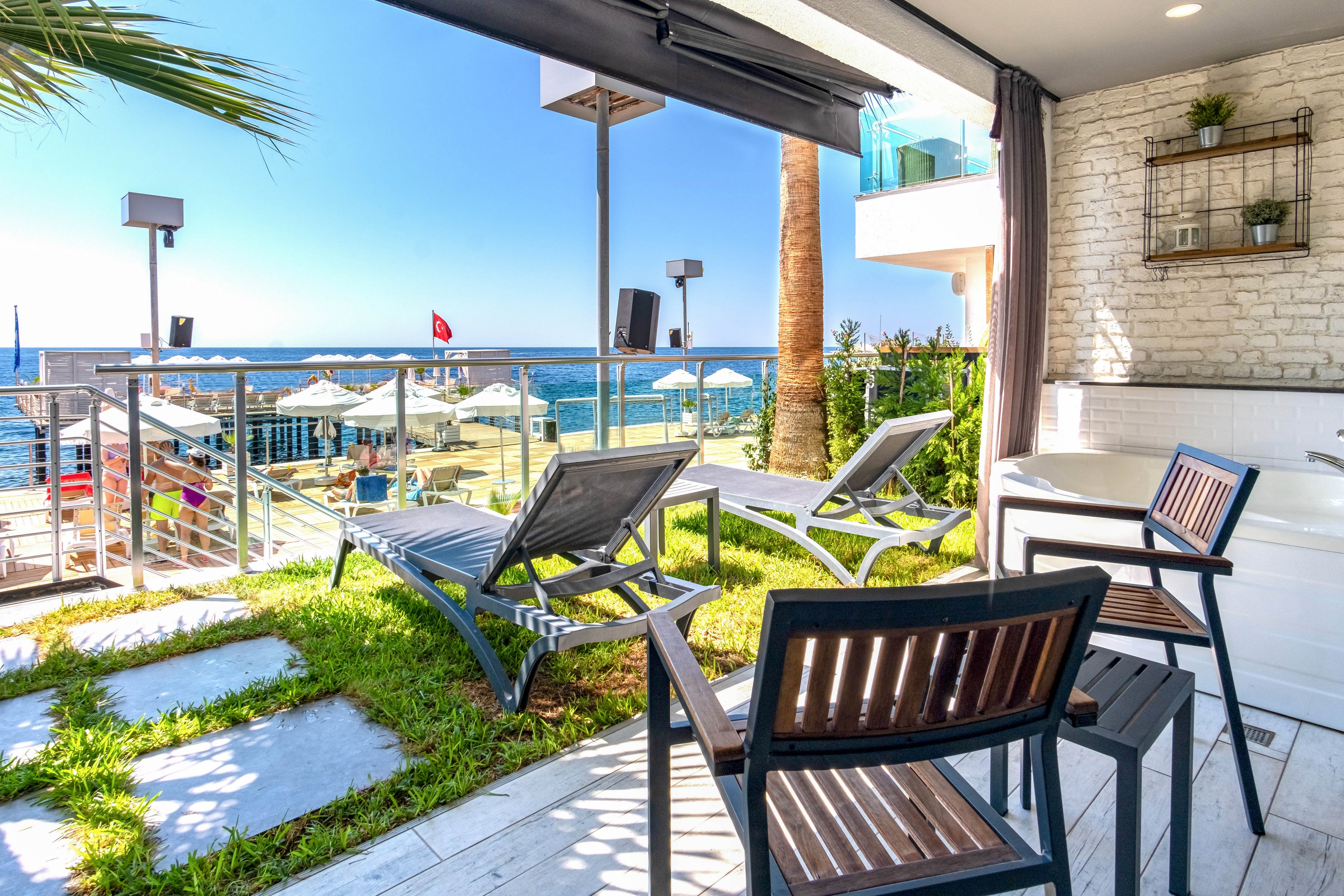 A contemporary terrace overlooks a sunny beachfront with clear blue water. Several lounge chairs are arranged on a small grassy area beside a sleek railing. In the distance, beach umbrellas and a pier with a red flag are visible. The bright daylight and clean design create a relaxing vacation atmosphere.