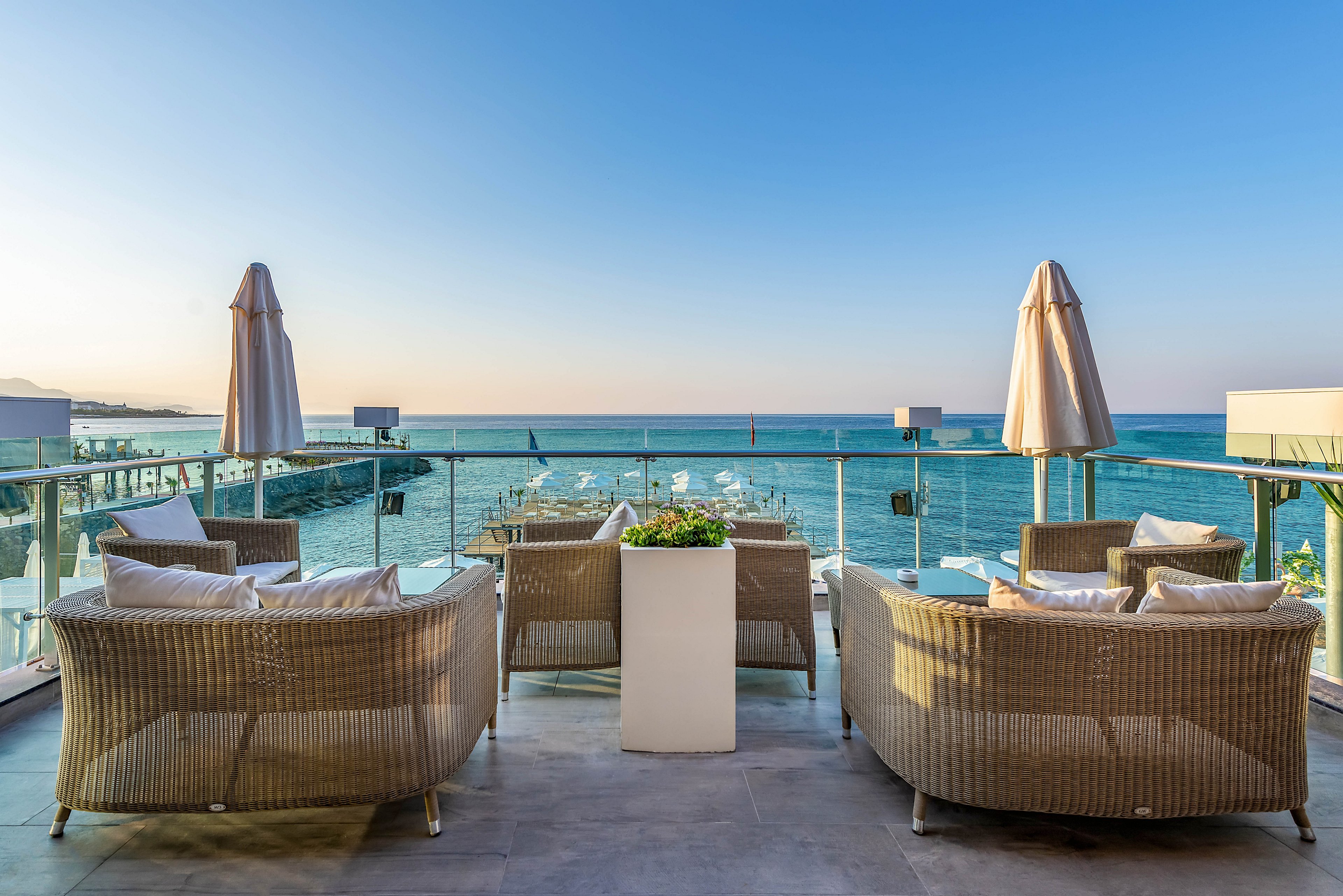 A contemporary outdoor lounge area overlooks a calm sea from an elevated terrace. Wicker armchairs with white cushions are arranged around a central planter, facing the ocean view. Glass railings, closed parasols, and a distant pier emphasize the coastal setting. The soft evening light and clear sky create a tranquil, luxurious atmosphere ideal for relaxation.