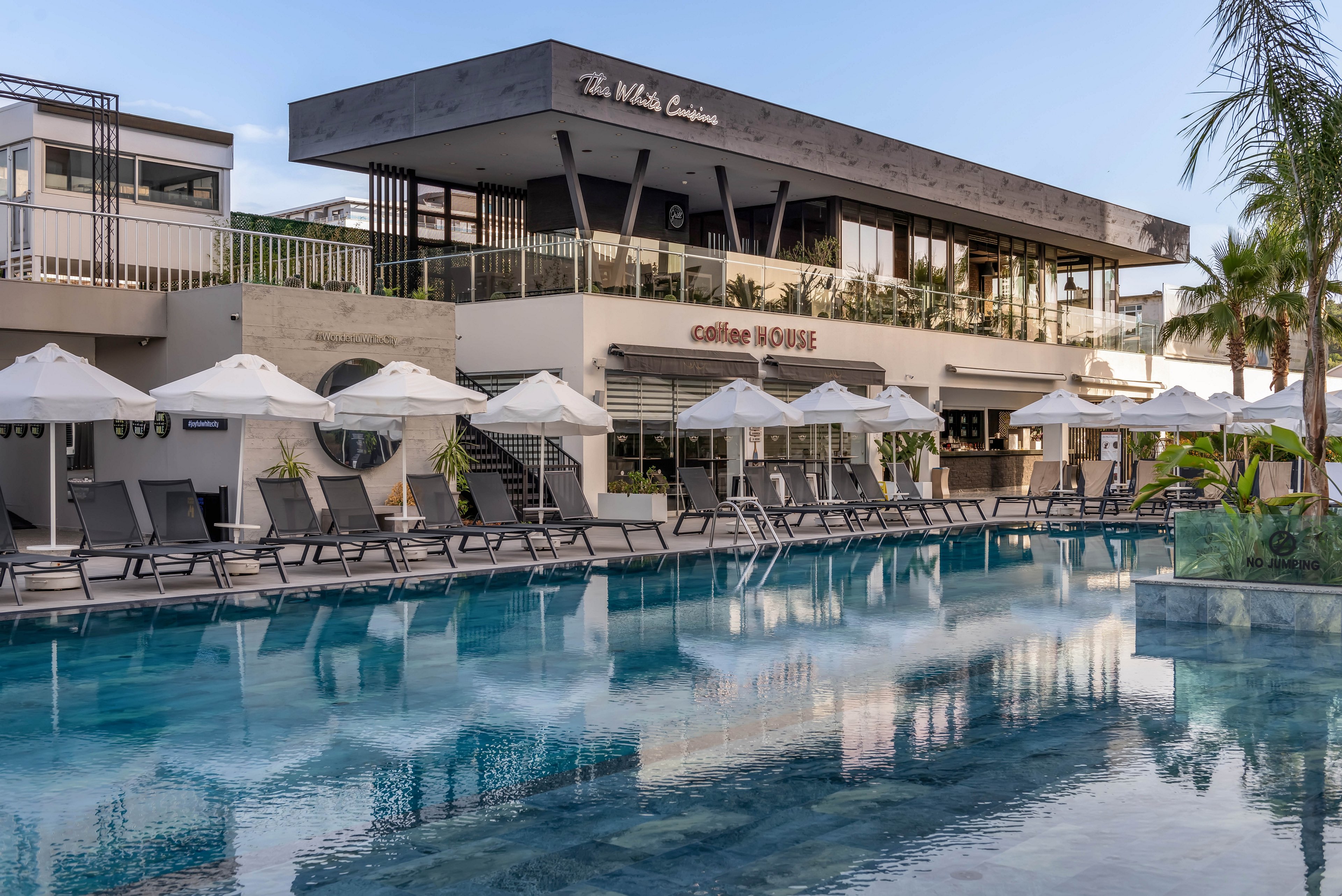A contemporary resort building overlooks a calm swimming pool lined with sun loungers and white umbrellas. The structure features clean architectural lines, glass railings, and an upper-level terrace. Reflections of the building and palm trees shimmer on the clear blue water. The scene suggests a relaxed vacation atmosphere in a warm, coastal destination.