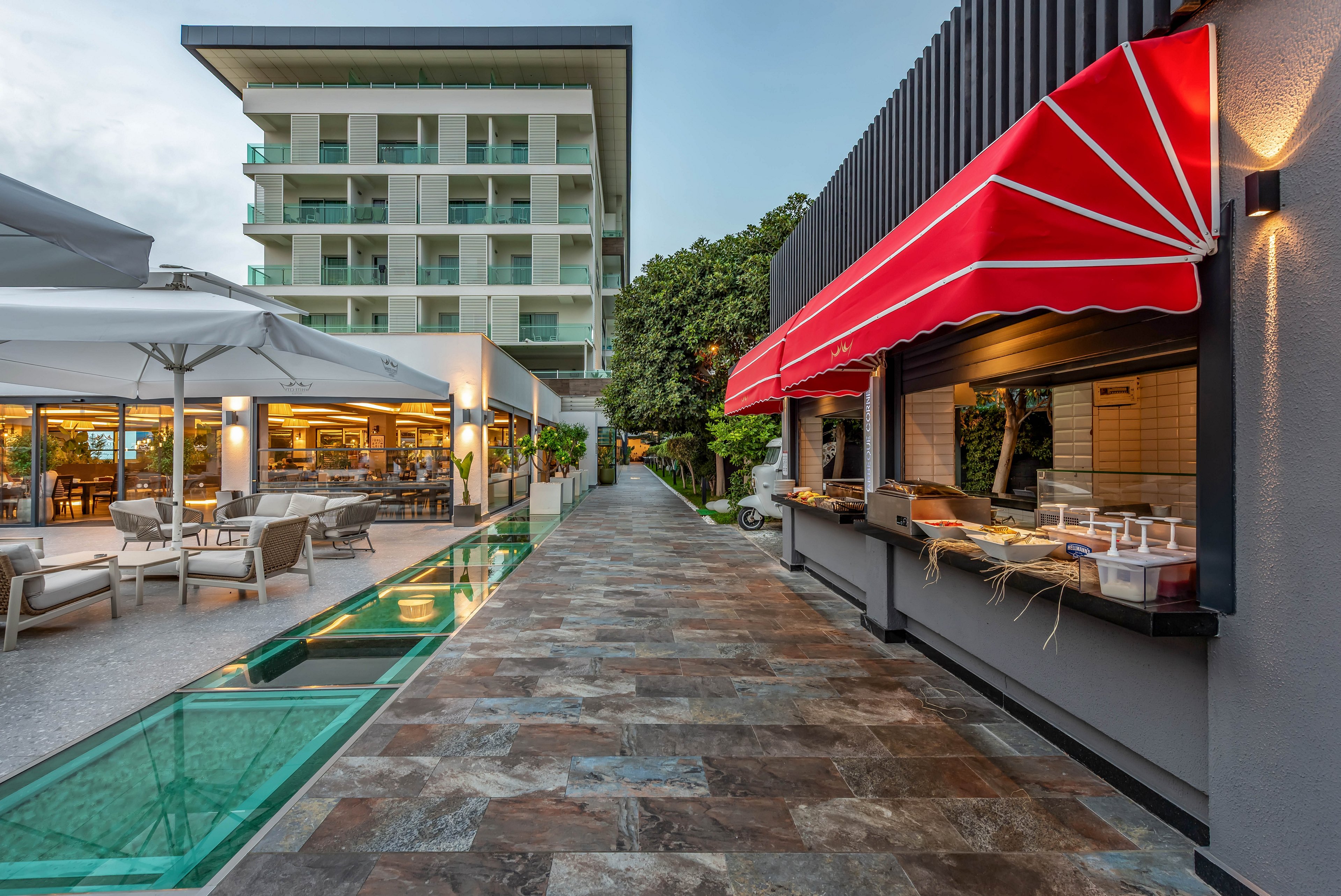 A contemporary hotel courtyard features an outdoor dining area beside a tall multi-story building. On the right, a food counter with a red striped awning displays dishes and utensils ready for service. To the left, guests sit at tables under large white umbrellas near a glass-sided water feature. The scene is captured at dusk, with warm interior lighting contrasting against the cool evening sky. The overall atmosphere feels relaxed and inviting for vacation or resort guests.