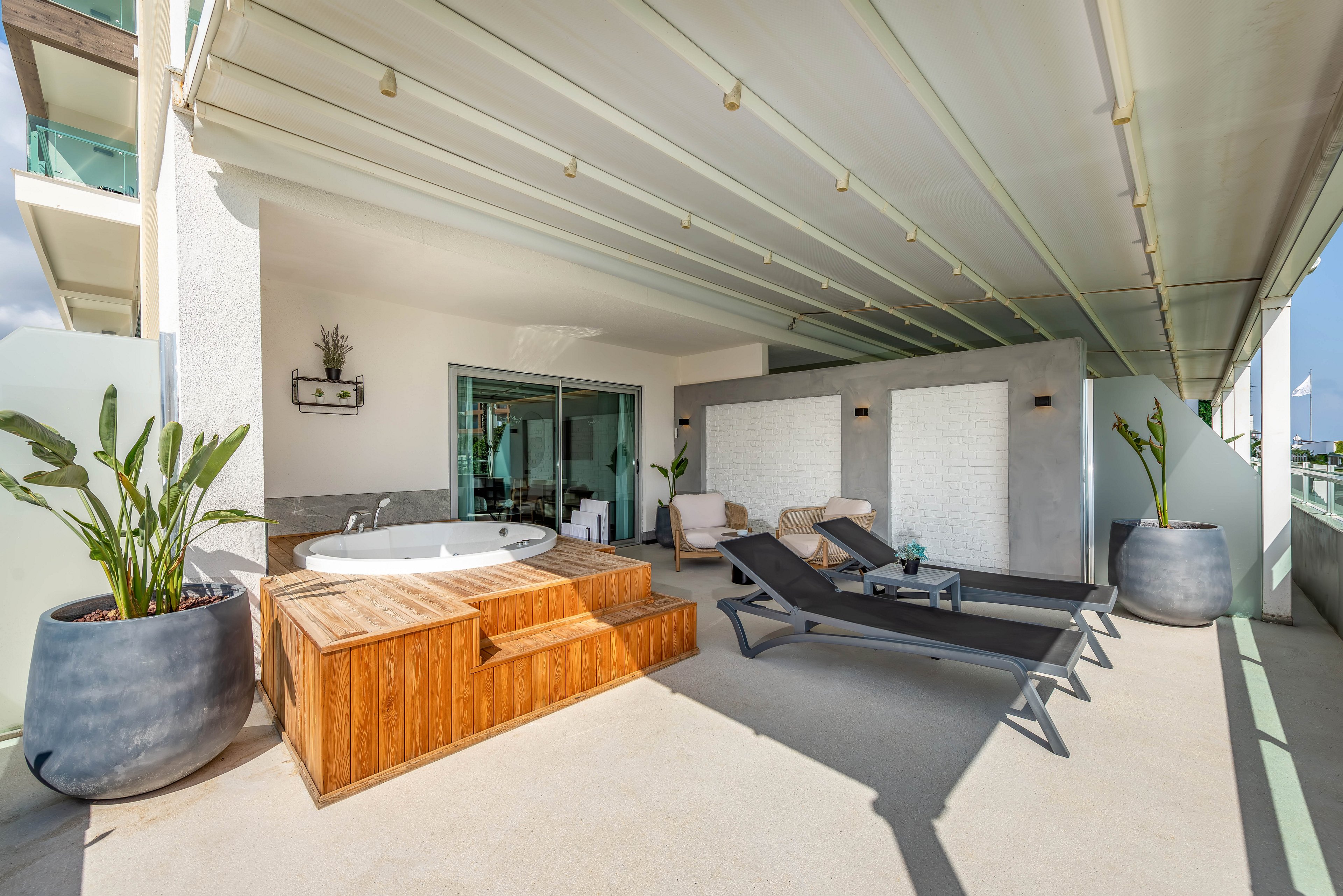 Spacious contemporary terrace features a built-in wooden jacuzzi as the focal point. Two black sun loungers and cushioned armchairs create a relaxing outdoor seating area. Large potted plants and frosted glass panels add privacy and greenery to the space. A retractable roof structure provides shade while sliding glass doors connect the terrace to the interior.