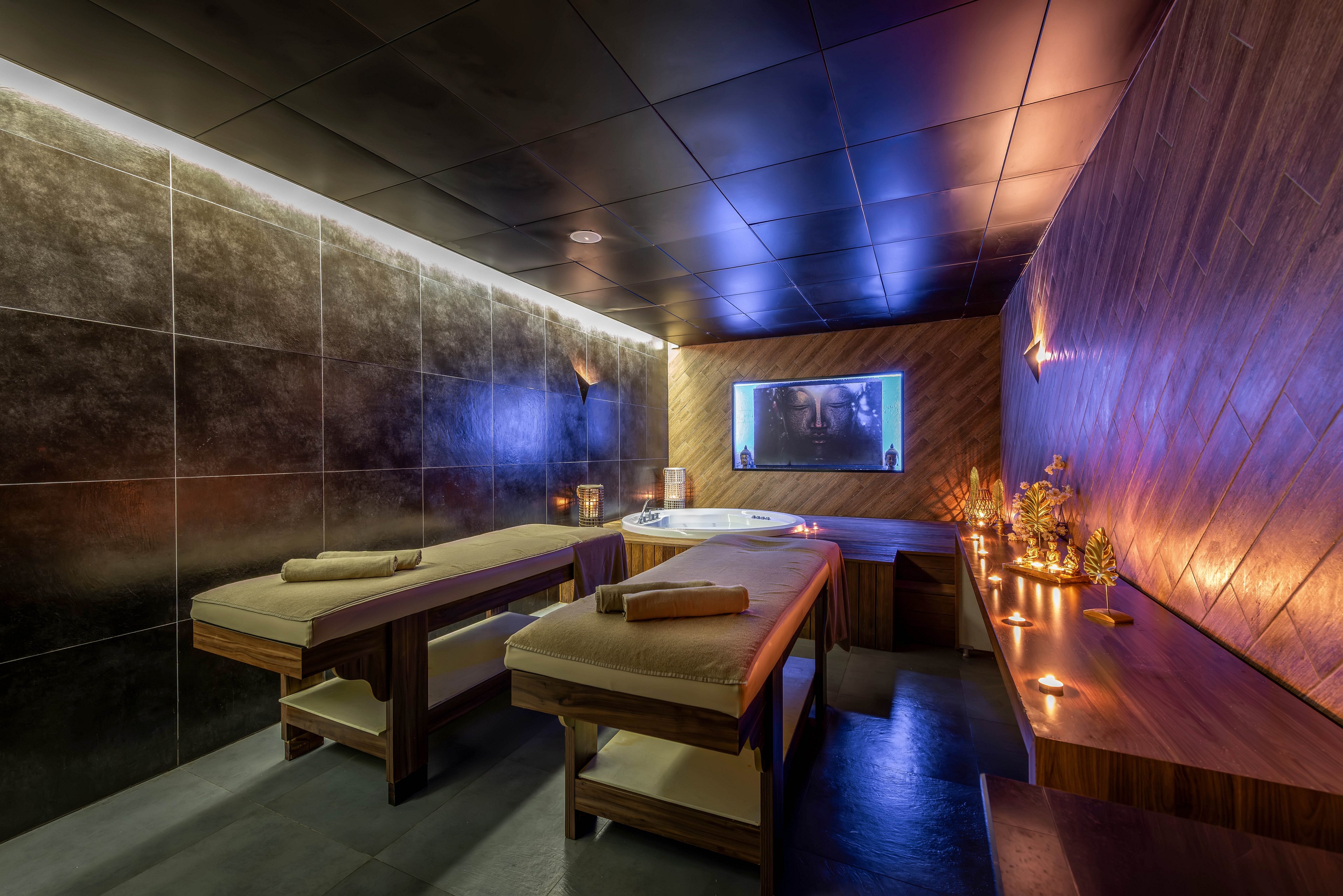 A modern spa treatment room features two massage tables neatly prepared with rolled towels. The space is softly illuminated by warm candlelight along a wooden counter and subtle recessed lighting in the ceiling. Dark tiled walls and sleek wooden surfaces create a contemporary, upscale atmosphere. A large screen on the far wall adds a relaxing visual focal point, enhancing the tranquil mood of the room.