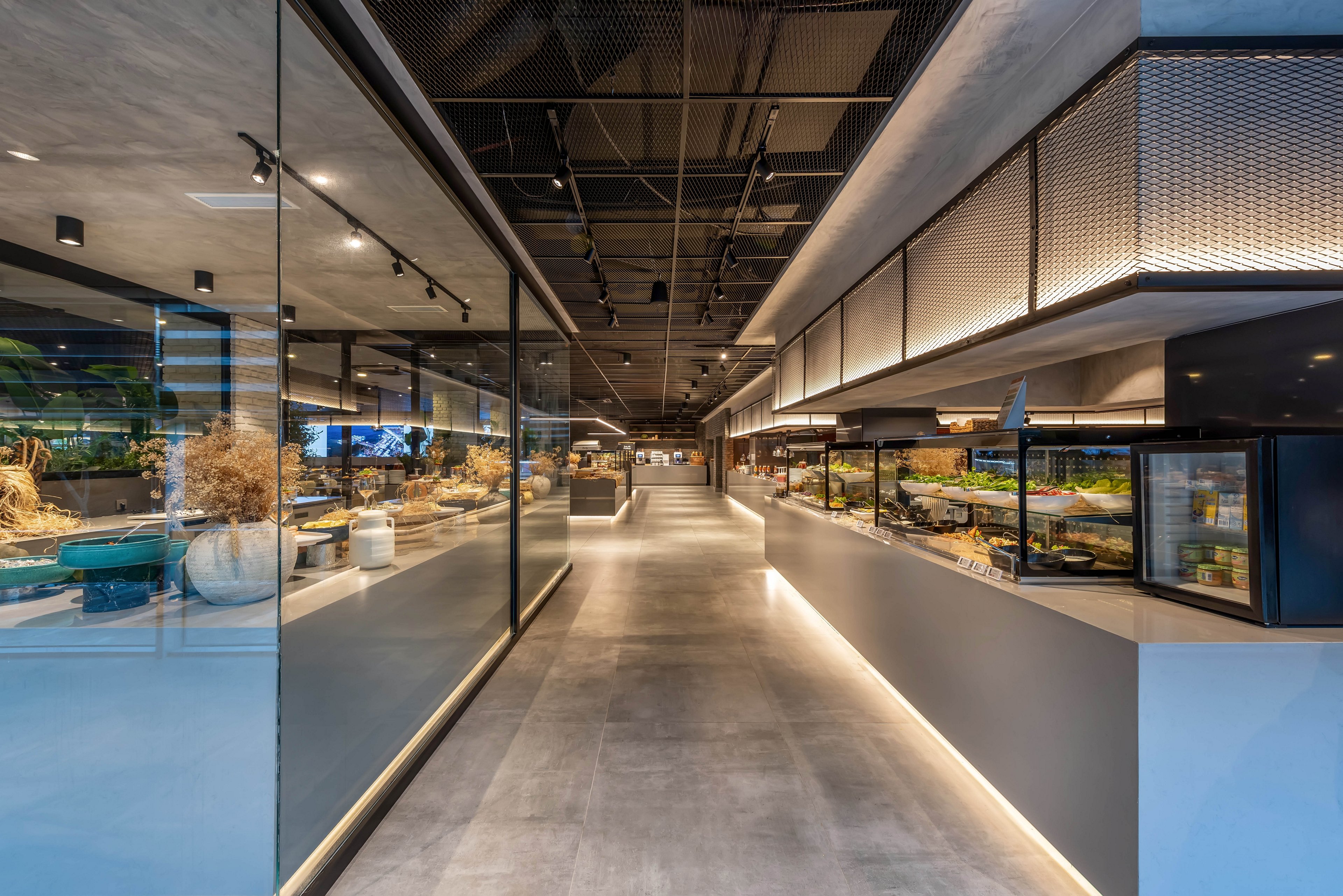 A sleek contemporary corridor features polished concrete floors and expansive glass walls. The space appears to be part of a modern commercial or retail interior with display counters on one side. Recessed lighting and exposed ceiling elements create a clean, industrial aesthetic. Decorative plants and curated objects add warmth and visual interest to the otherwise minimalist design.