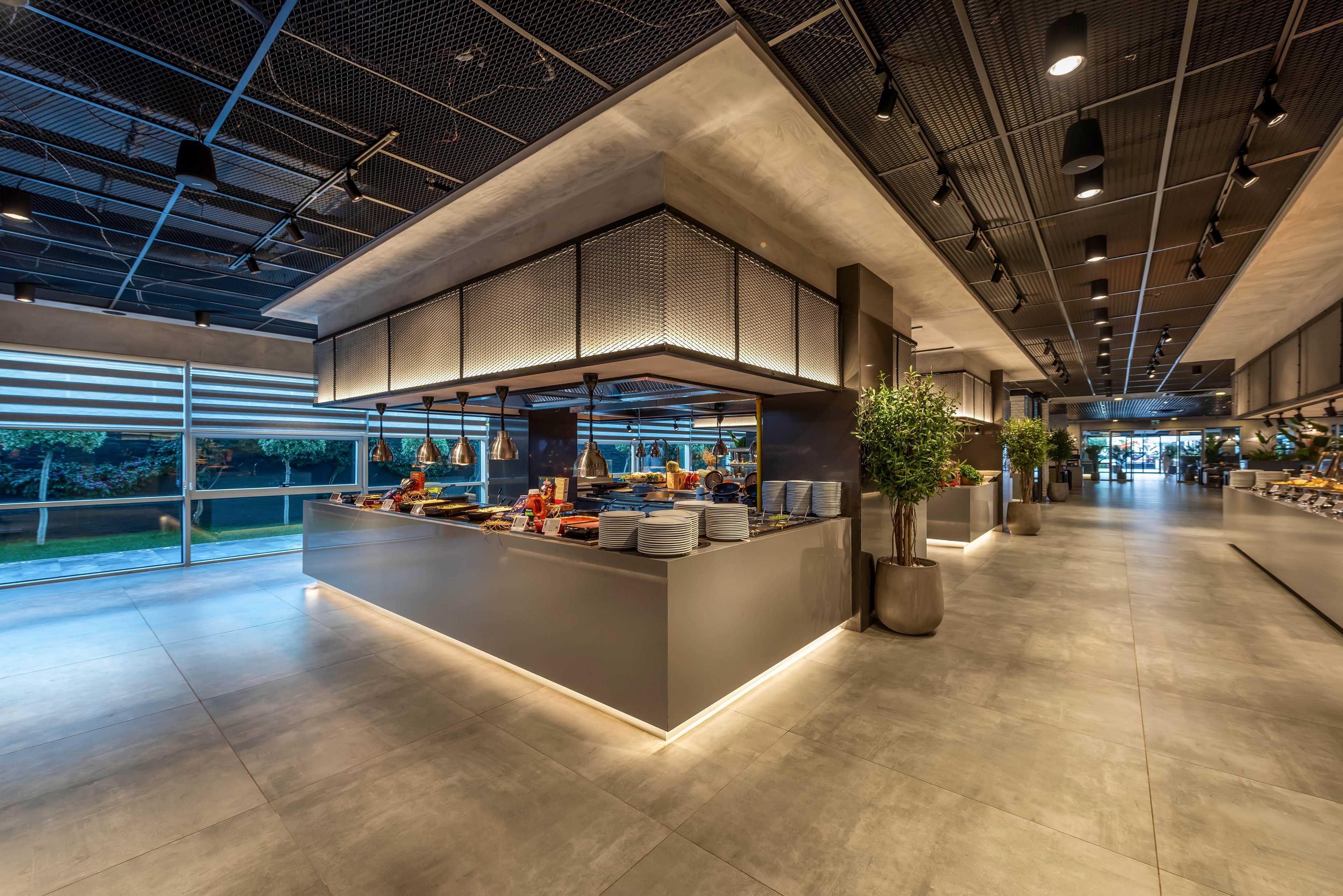 A contemporary buffet station stands in the center of a spacious restaurant interior. Sleek grey counters display plates, bowls, and an assortment of food under warm lighting. Large floor-to-ceiling windows reveal greenery outside, adding natural contrast to the industrial ceiling and polished floor. The scene feels clean, organized, and ready for guests in a high-end dining environment.
