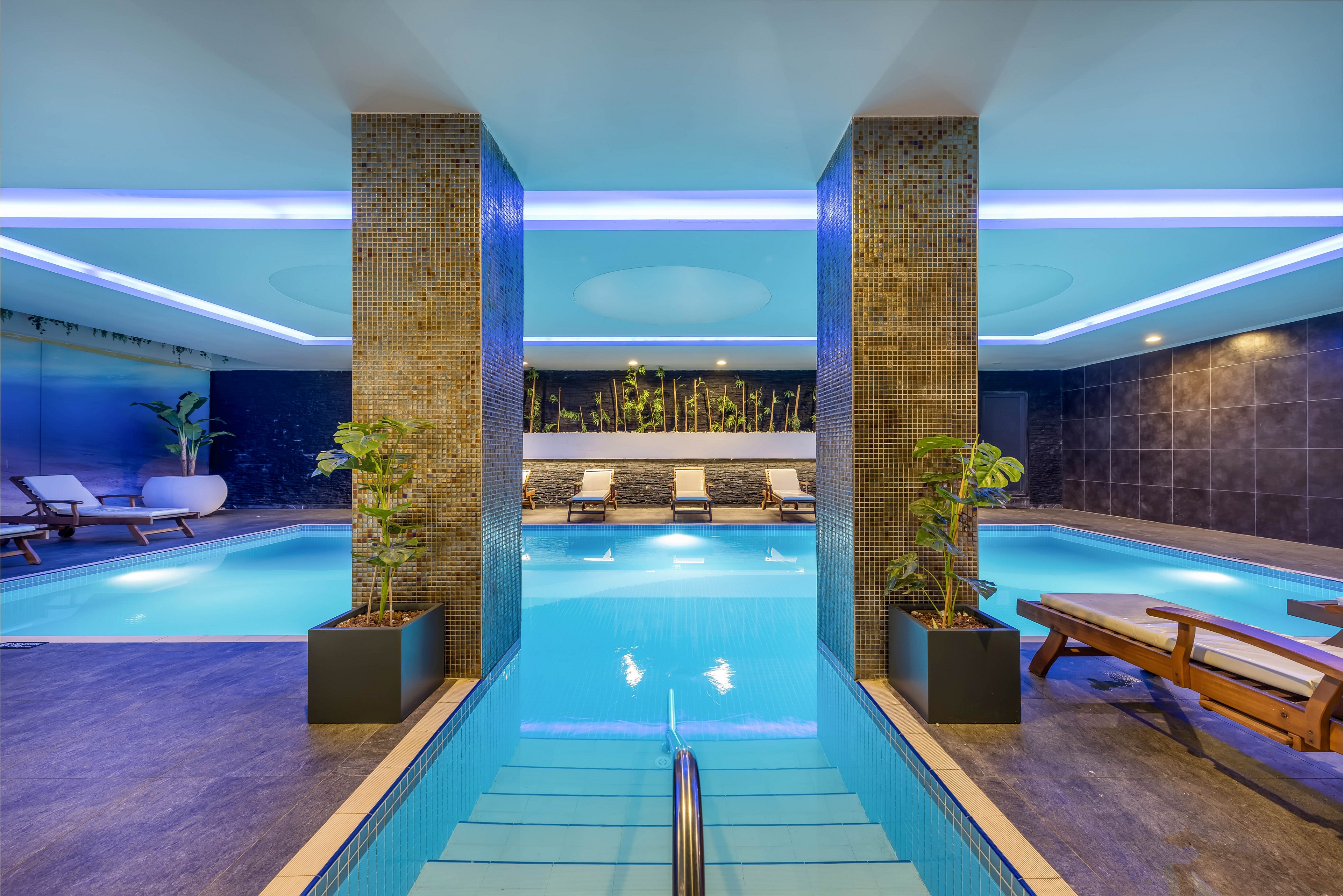 A contemporary indoor swimming pool area features a central walkway leading into clear blue water. Two tiled columns with potted plants divide the pool, while lounge chairs line the sides for relaxation. Soft blue and white ceiling lights create a calm, upscale atmosphere. Dark wall tiles and decorative greenery at the far end enhance the spa-like, tranquil setting.