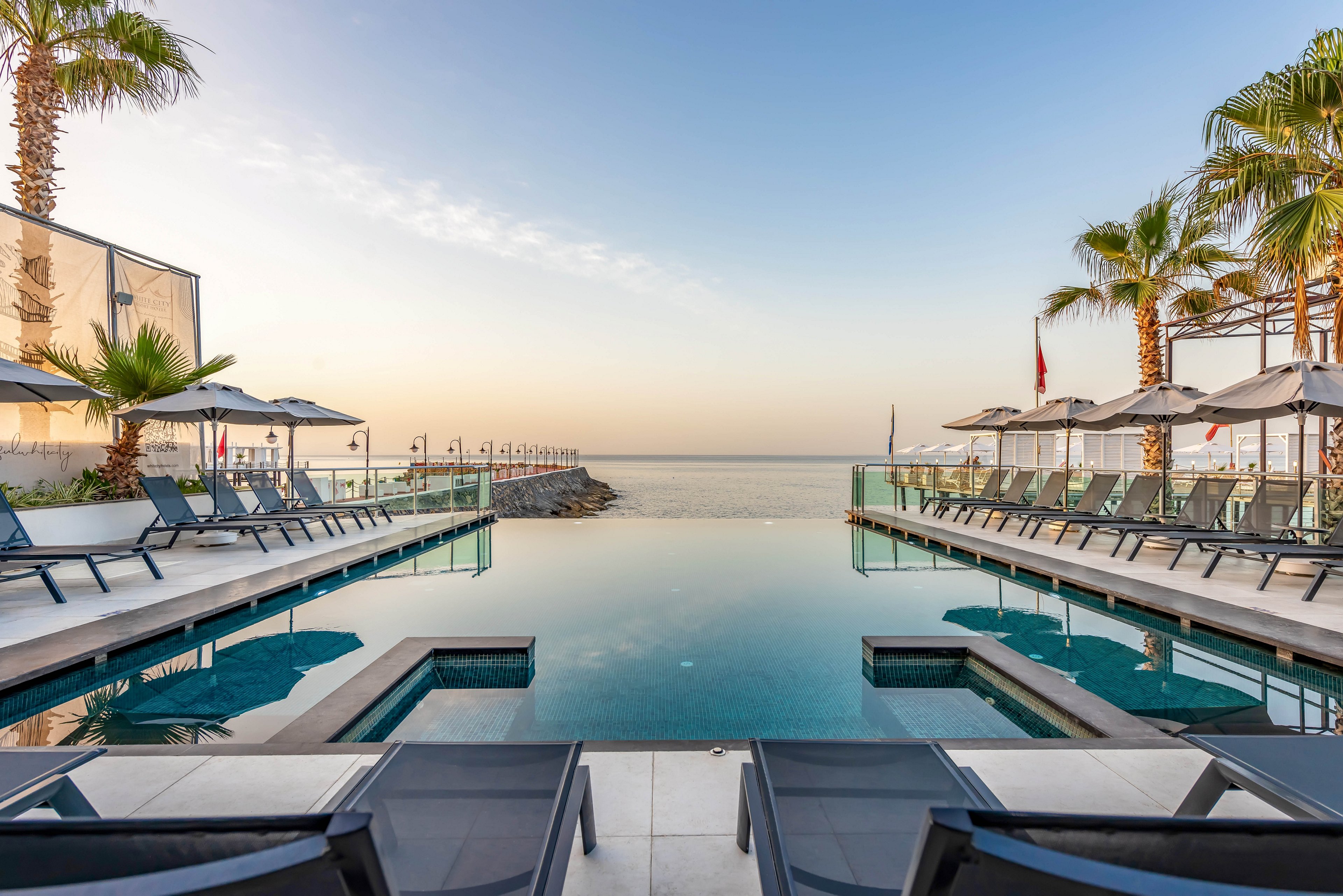 A modern infinity pool stretches toward a calm ocean horizon at sunrise or sunset. Sun loungers and umbrellas line both sides of the pool deck, creating a symmetrical resort setting. Tall palm trees frame the scene, adding a tropical atmosphere. The clear sky and soft warm light enhance the serene, luxurious vacation mood.