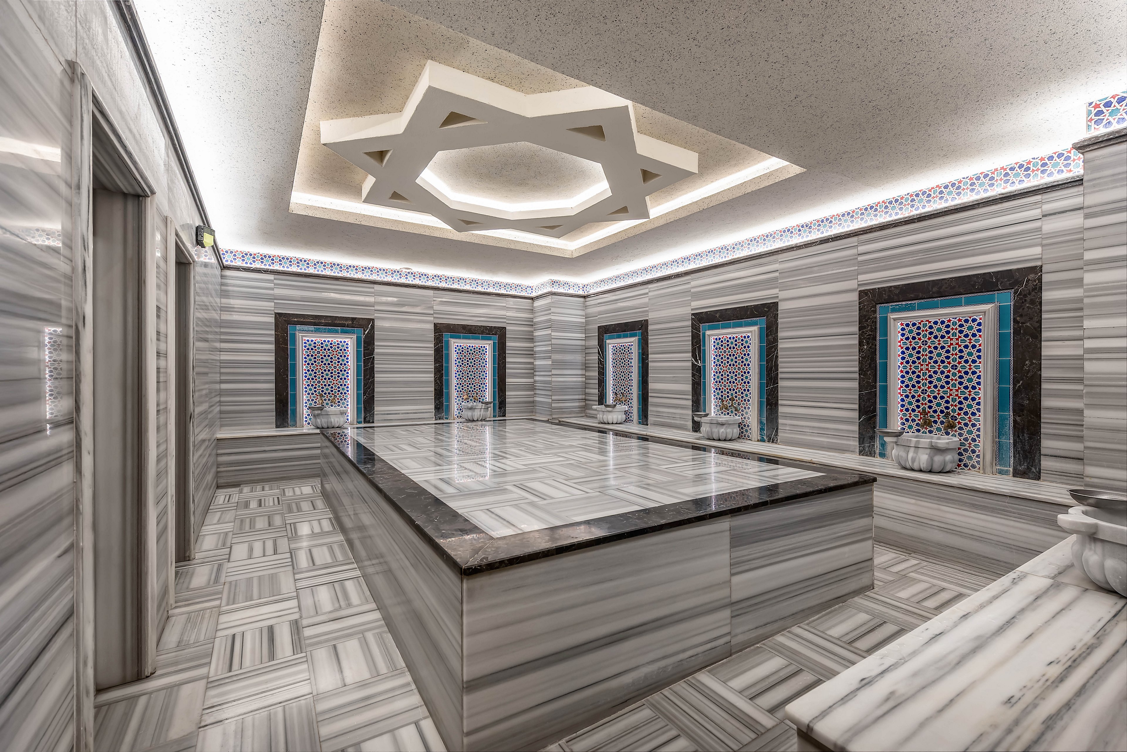 Spacious marble-clad Turkish hammam with a large central platform dominates the scene. The room features striped gray stone walls and floors, accented by geometric tile panels around the basins. A striking star-shaped ceiling light adds a contemporary touch to the traditional bathhouse architecture. The space appears clean, serene, and ready for spa or wellness rituals.