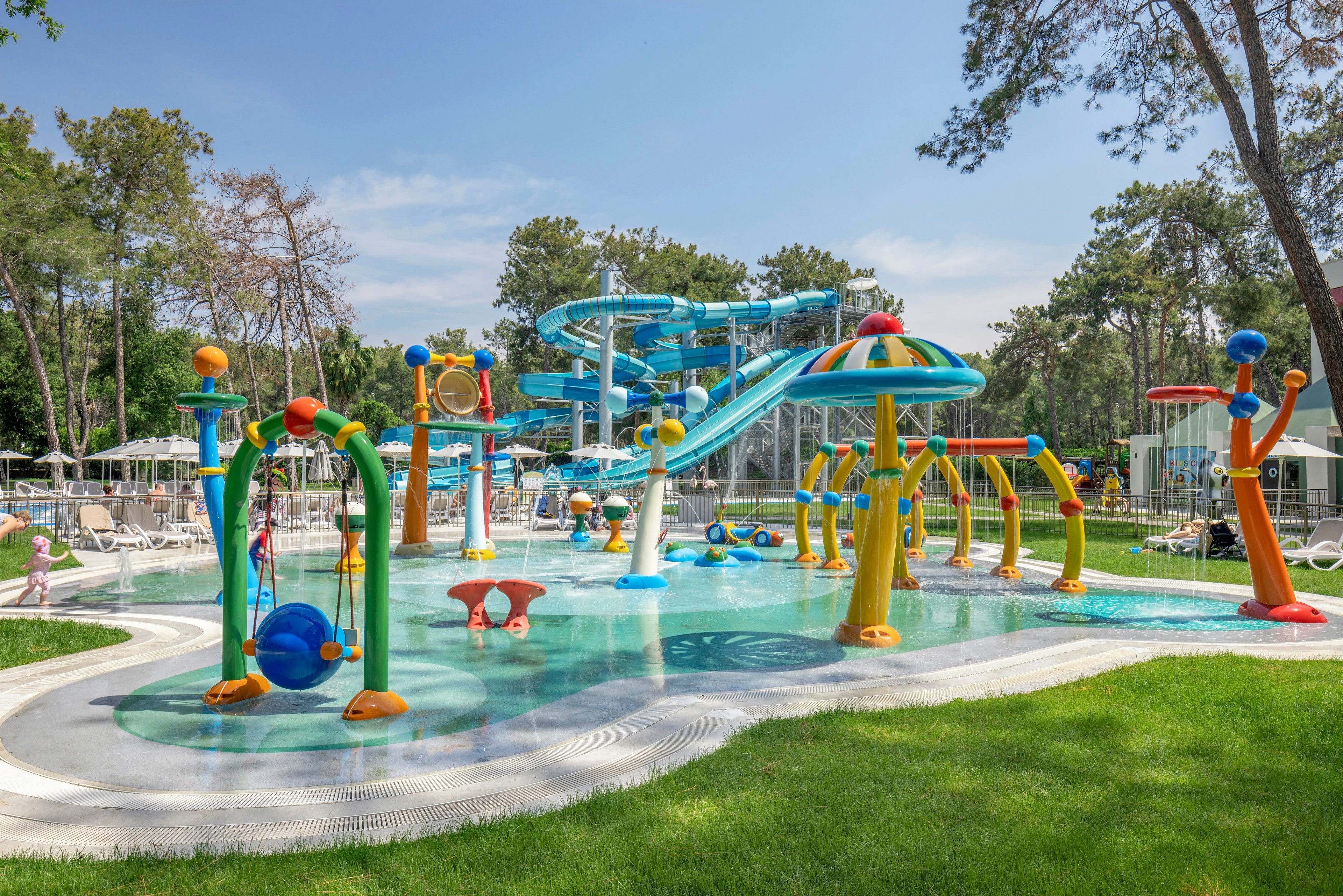 A vibrant outdoor water park features multiple colorful splash structures and fountains in a shallow pool. In the background, large twisting water slides rise above the play area, adding excitement to the scene. Lounge chairs and umbrellas surround the pool, indicating a family-friendly recreational setting. Tall trees and green grass frame the park, creating a relaxed holiday atmosphere under a clear blue sky.
