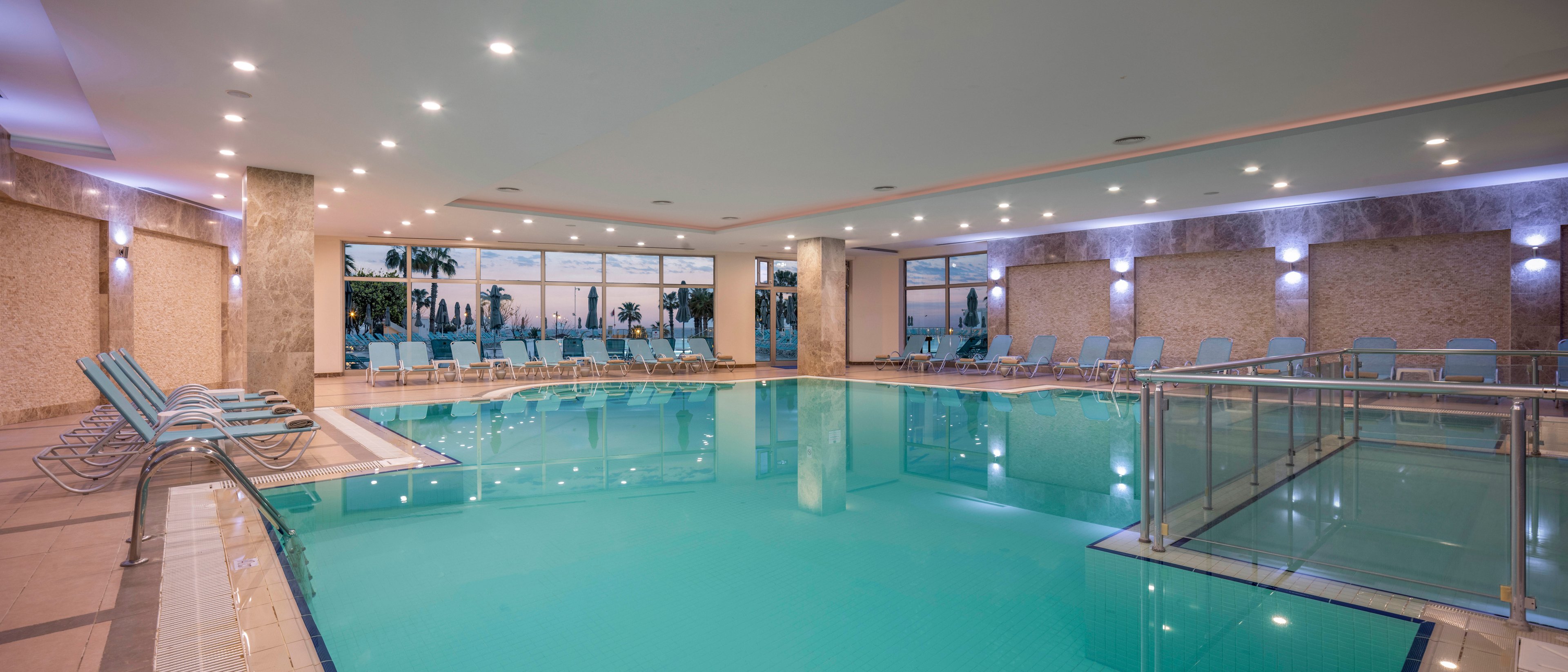 Spacious indoor swimming pool with calm turquoise water is surrounded by light blue lounge chairs. Large floor-to-ceiling windows reveal an evening sky and silhouettes of palm trees outside. Recessed ceiling lights and wall sconces create a soft, inviting glow on the stone walls. A stainless steel handrail and steps provide easy access to the pool, emphasizing comfort and relaxation.