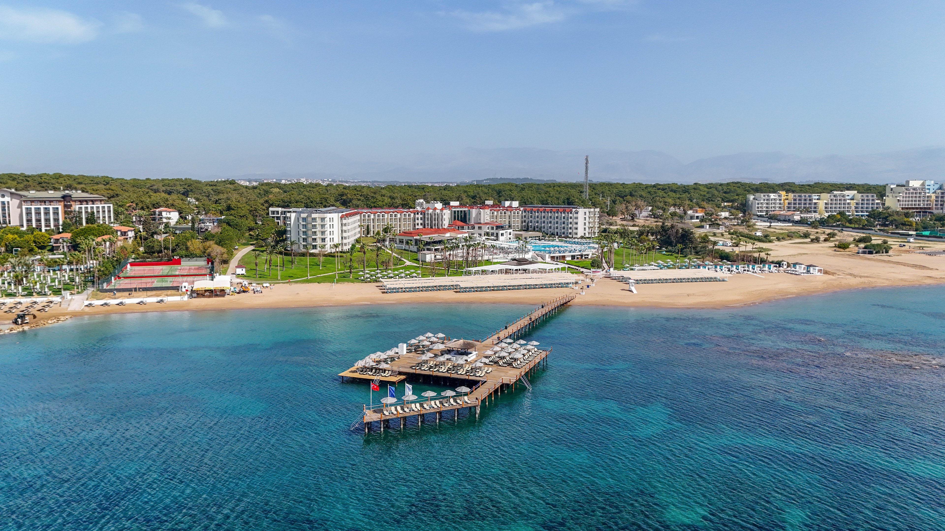 A large beachfront resort complex stretches along a sandy coastline beside clear blue water. An extended wooden pier with sun loungers and umbrellas reaches out into the calm sea. Several hotel buildings are surrounded by greenery and palm-lined walkways. The scene suggests a tranquil holiday destination with ample beach and waterfront amenities.