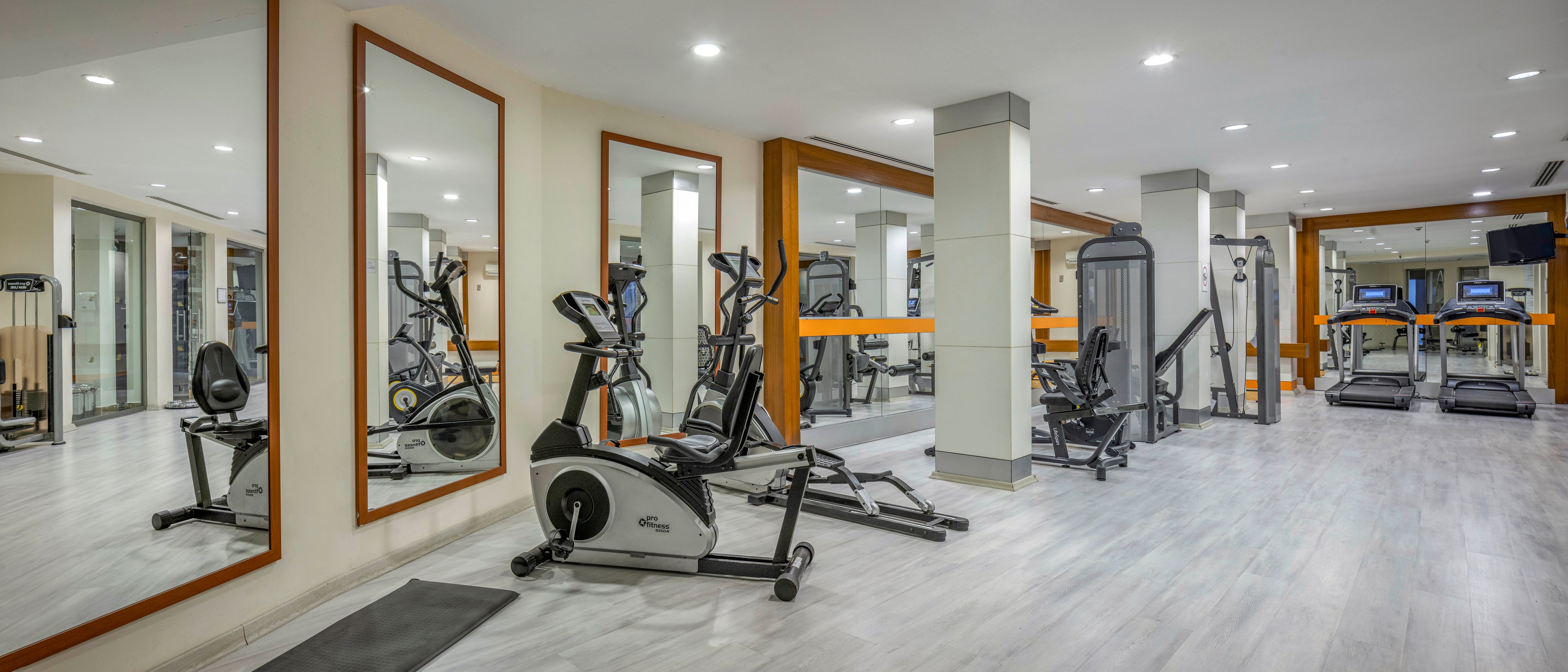 A spacious, modern gym features various exercise machines including treadmills, stationary bikes, and weight equipment. Large wall mirrors visually expand the room and reflect the workout area. Bright overhead lighting and light-colored flooring create a clean, contemporary atmosphere. The space appears organized and ready for guests to use for cardio and strength training.