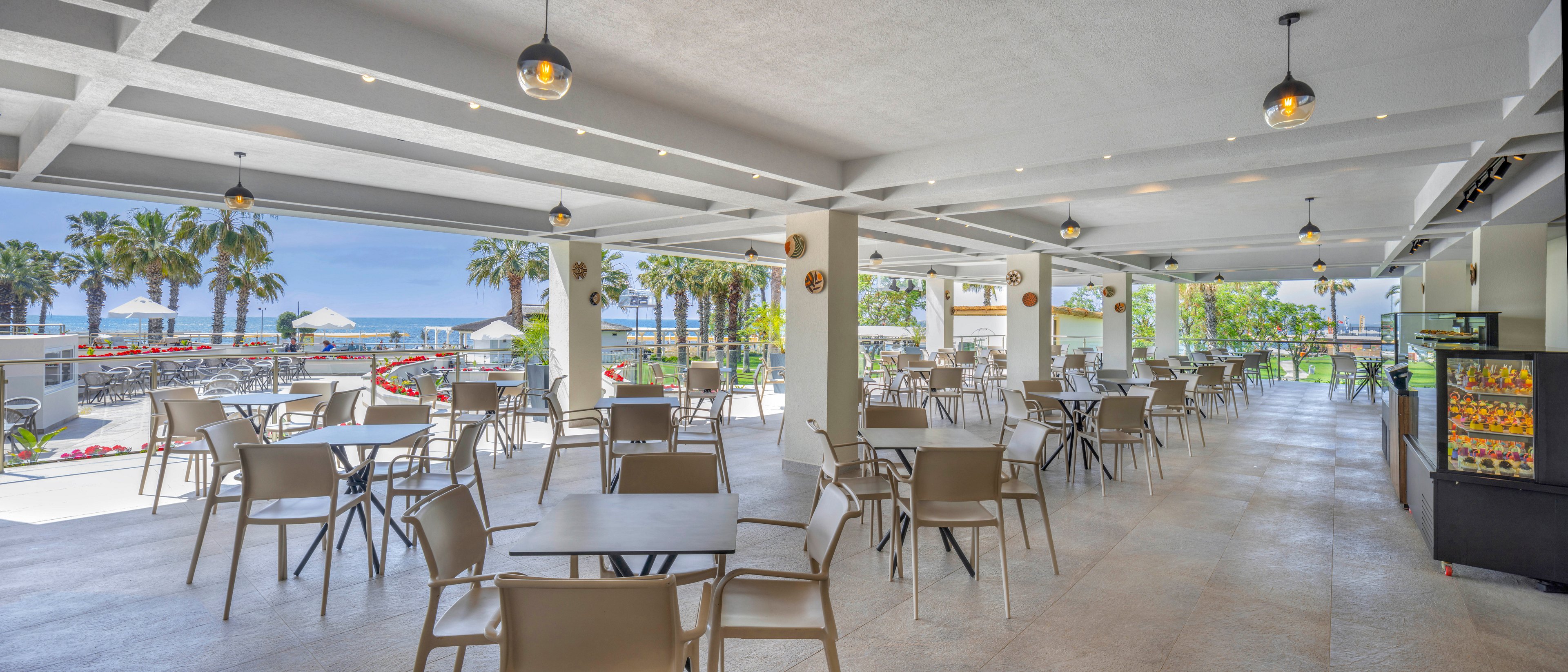 An open-air café with many empty tables and chairs overlooks a sunny beachfront. The covered terrace features white beams, hanging pendant lights, and a tiled floor. In the background, palm trees, umbrellas, and the blue sea create a relaxed vacation atmosphere. A refrigerated display case with drinks and snacks stands on the right side of the image.