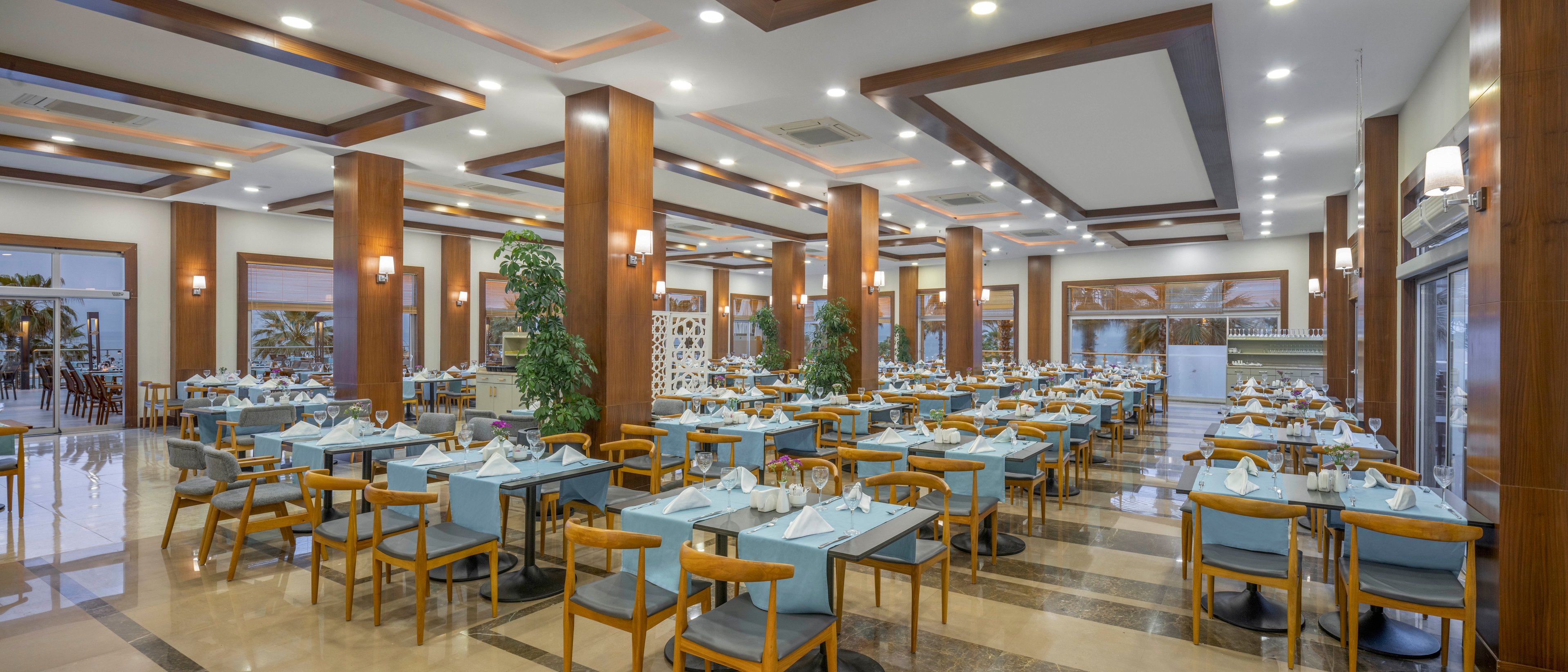 A large, modern restaurant dining room is set with neatly arranged tables and chairs. Light blue table linens and wooden furniture create a clean, contemporary look. Tall wooden columns, recessed ceiling lights, and wall sconces brighten the space. The room appears ready for guests, with folded napkins, glassware, and cutlery placed at each setting.