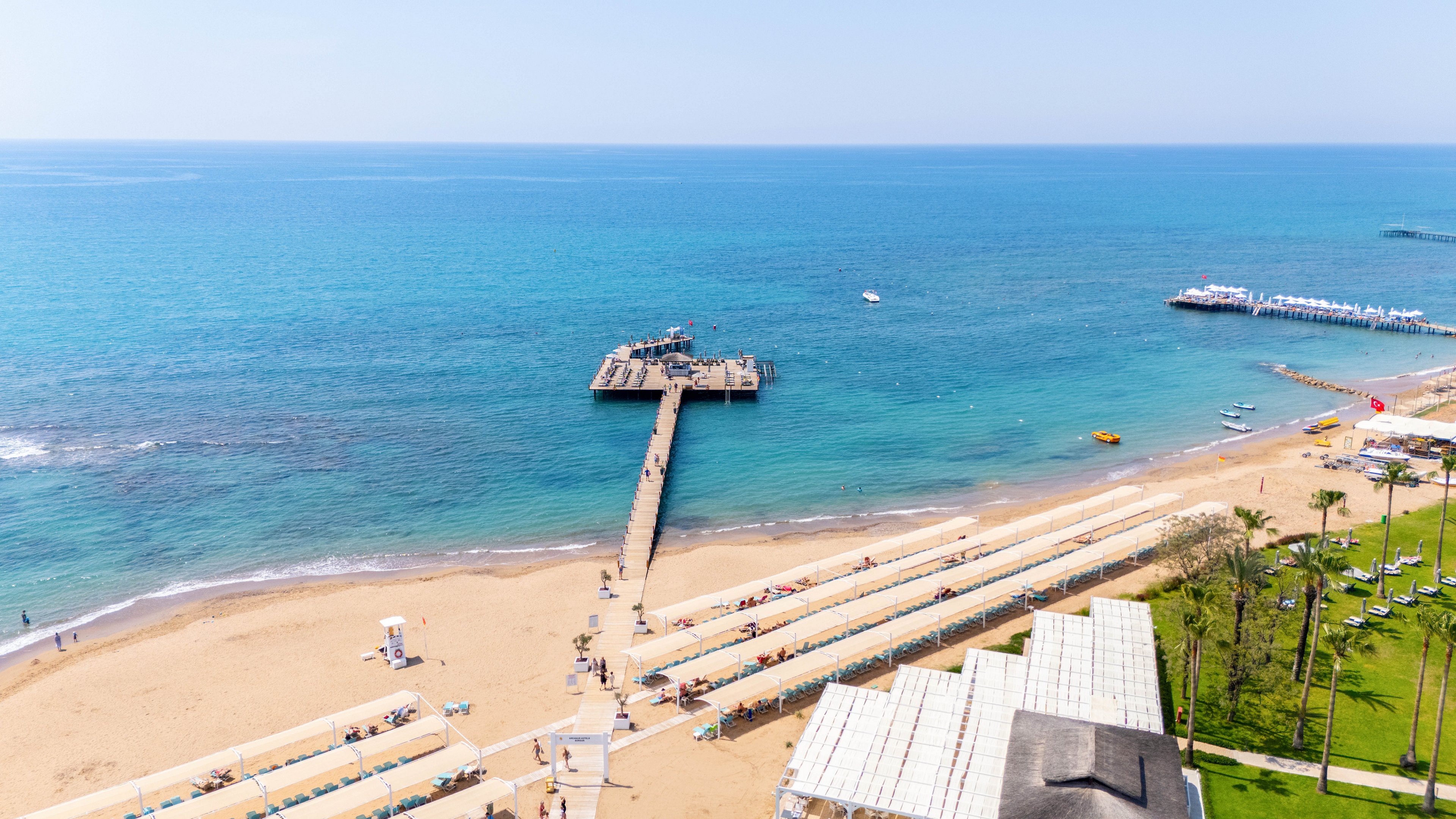 A wide sandy beach stretches along a bright turquoise sea at a coastal resort. A long wooden pier extends out into the calm water, with people walking and relaxing along its length. Neatly arranged rows of sun loungers and umbrellas line the shore, suggesting a popular vacation destination. Green lawns and palm trees frame the beachfront, creating a relaxed, summery atmosphere under a clear blue sky.