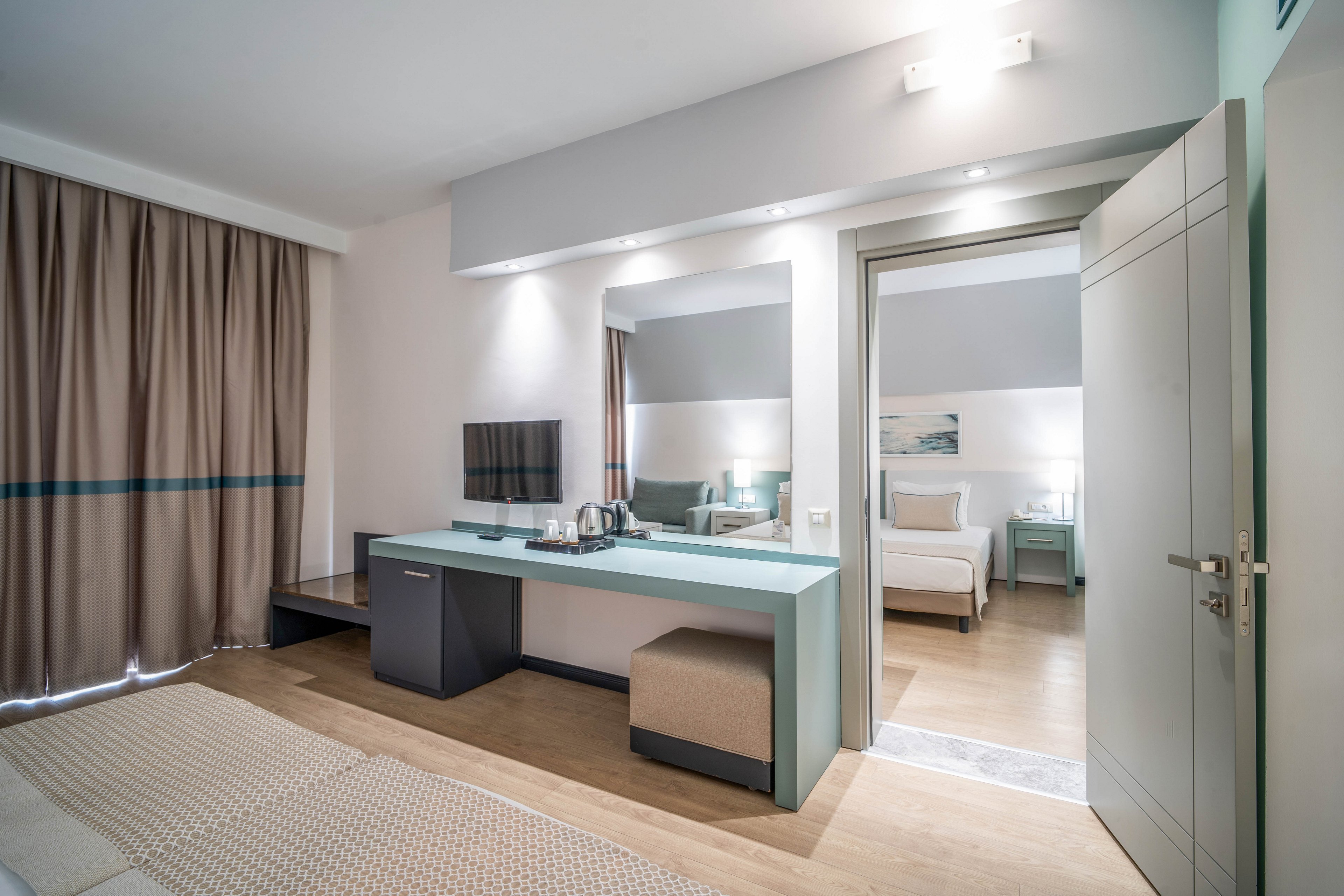 Contemporary hotel room interior featuring a desk with a flat-screen TV and hospitality tray. A large mirror and soft neutral furnishings create a clean, minimalist look. The open door reveals an adjoining bedroom with a neatly made bed and bedside table. Warm wood flooring and soft lighting contribute to a calm, inviting atmosphere.