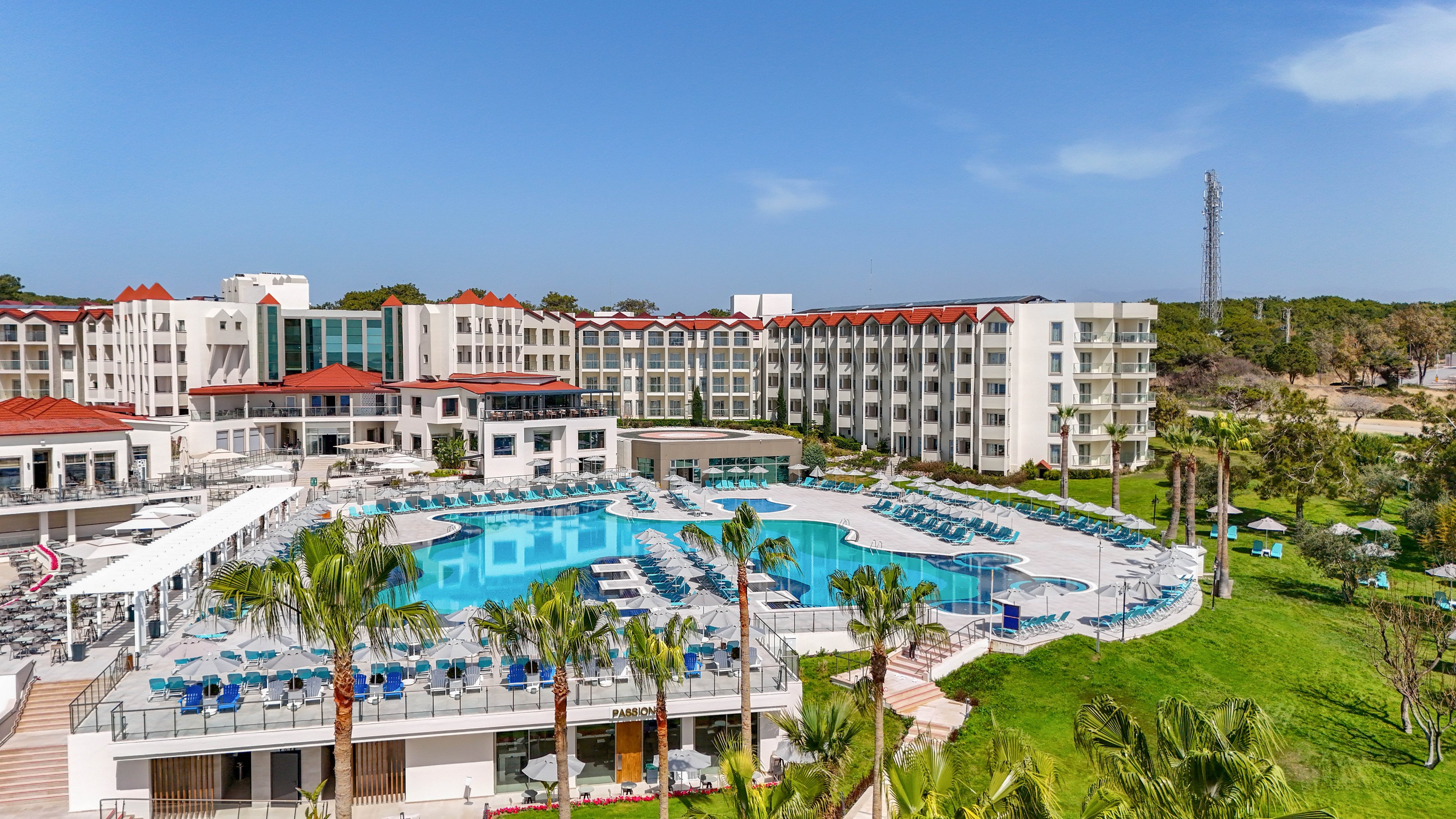 A large resort hotel complex features multiple white buildings with red roofs surrounding a central outdoor swimming pool. Sun loungers and umbrellas are neatly arranged around the poolside terraces, suggesting a relaxing vacation setting. Lush green lawns, palm trees, and landscaped gardens extend from the pool area toward the edge of the property. The scene is captured on a bright, clear day with blue sky, emphasizing a warm and inviting holiday atmosphere.