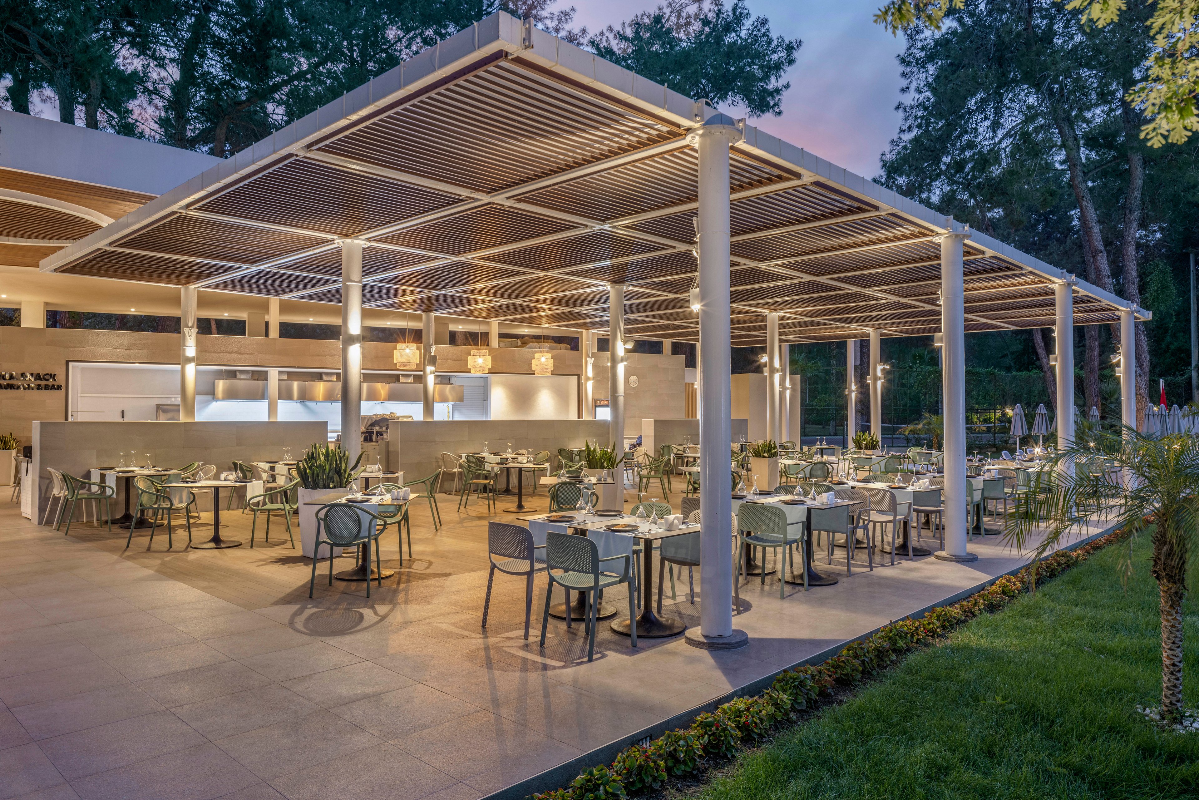 A spacious open-air restaurant terrace is set with neatly arranged tables and chairs under a modern pergola structure. Warm ambient lighting from ceiling fixtures and hanging lamps creates an inviting atmosphere as evening approaches. The clean architectural lines and neutral tones contrast with the surrounding greenery and manicured lawn. The scene suggests a contemporary dining experience in a relaxed outdoor setting.