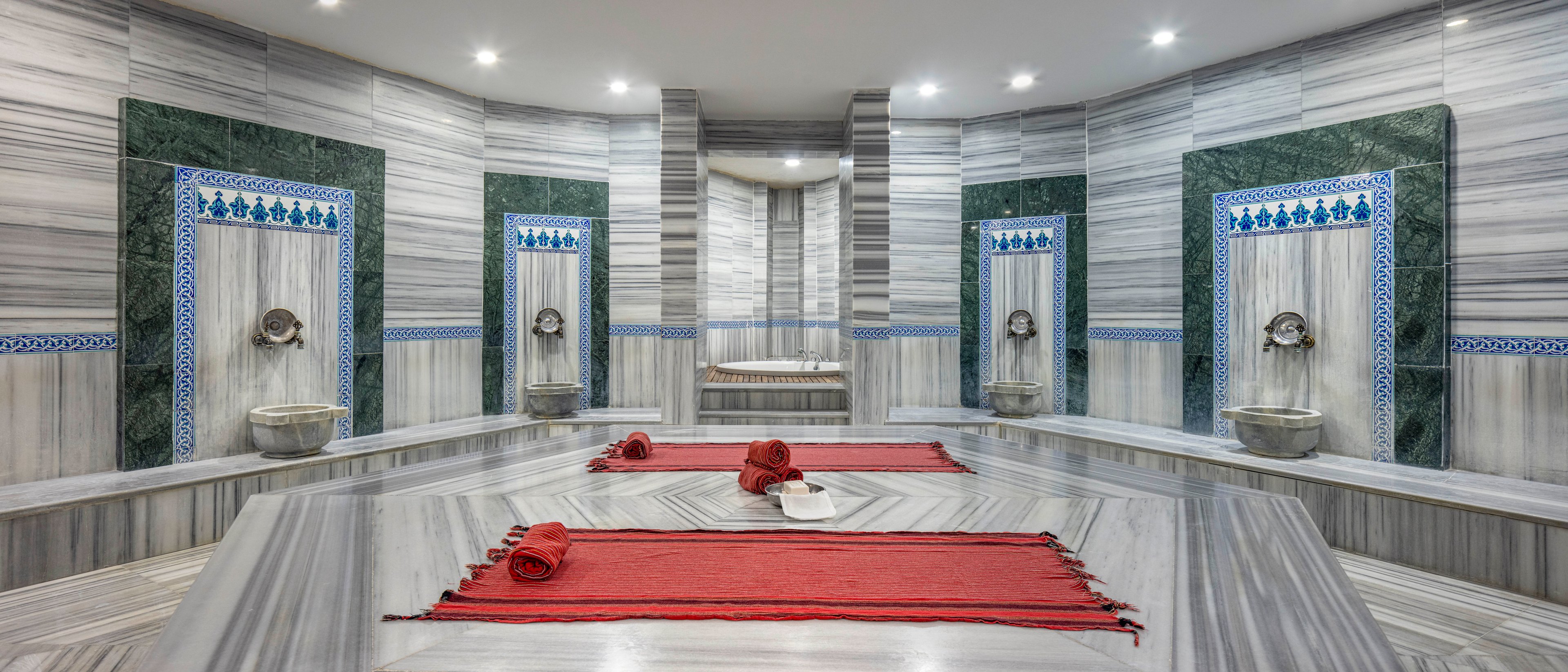 Spacious marble Turkish bath interior with a central heated platform is shown from a symmetrical perspective. Stone basins with metal faucets line the walls, framed by decorative green and blue tiled panels. Red towels are neatly rolled and laid on the central marble surface, adding a vivid contrast to the grey stone. Recessed ceiling lights create a bright, serene spa atmosphere.