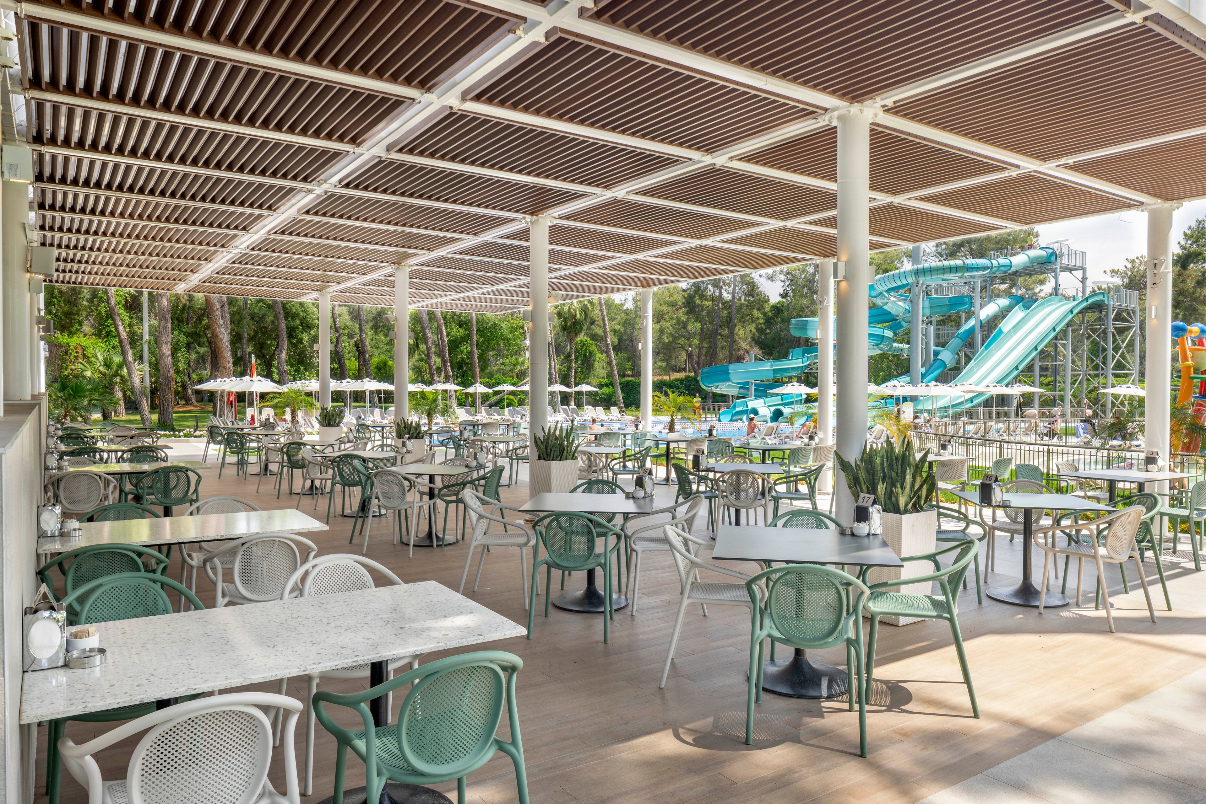 An open-air cafe terrace features neatly arranged tables and chairs under a slatted roof. The seating area is mostly empty, creating a calm and orderly atmosphere. In the background, a colorful water park with large blue water slides and pools is visible among green trees. The scene suggests a resort or leisure complex designed for outdoor dining and family recreation.