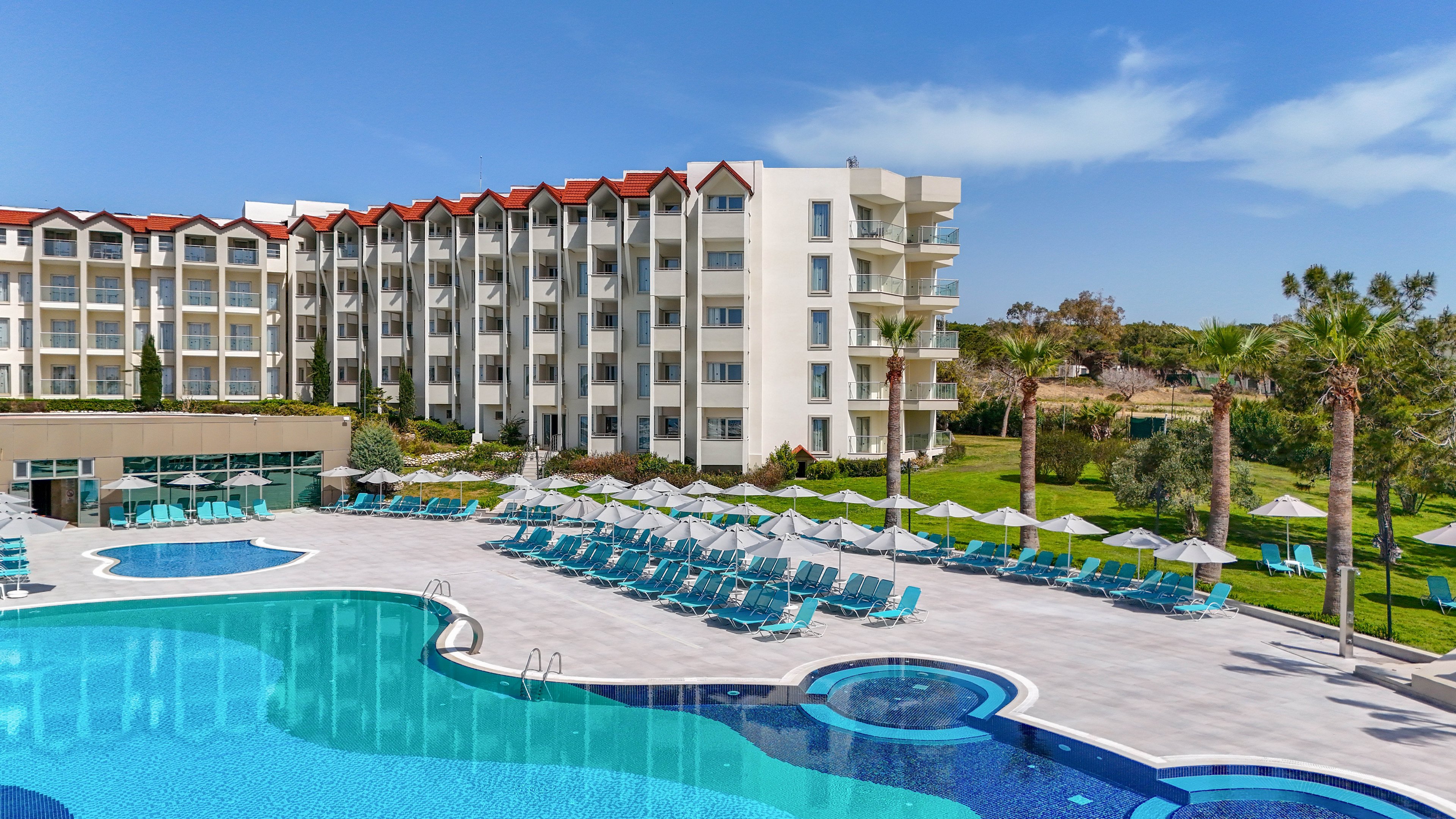 A large resort hotel stands beside a spacious outdoor swimming pool under a clear blue sky. Numerous turquoise sun loungers and white umbrellas line the pool deck, creating a relaxed vacation atmosphere. The multi-story building features balconies overlooking the pool and landscaped green lawns with palm trees. The scene suggests a sunny, warm-weather destination ideal for leisure and holidays.