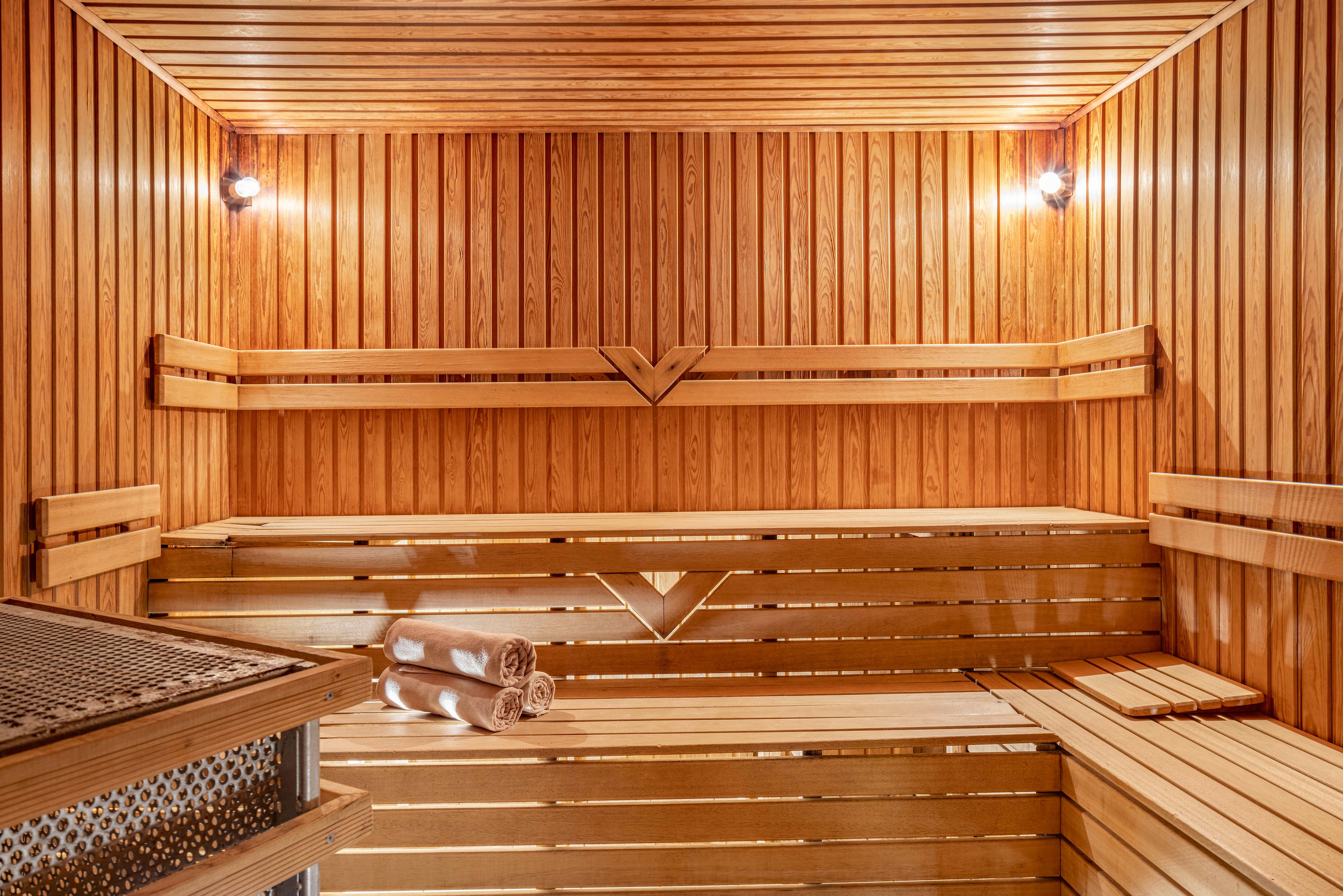 A warm, empty wooden sauna interior is shown with tiered benches along the walls. Soft lighting from two wall-mounted lamps highlights the natural wood grain and slatted construction. Rolled towels rest neatly on one of the lower benches near the heater area. The scene conveys a clean, inviting spa environment ready for relaxation.