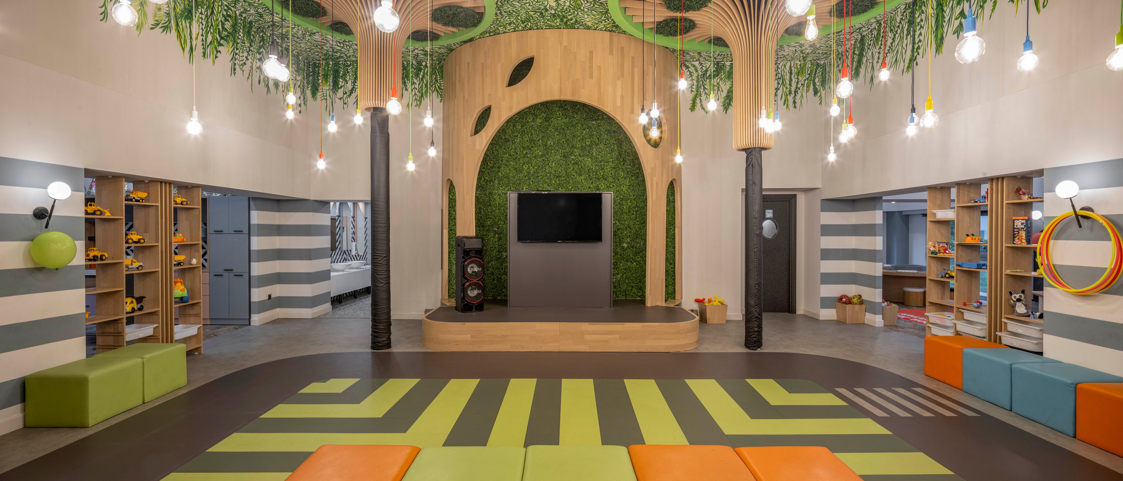 A spacious children's playroom features a central stage area with a large wall-mounted screen framed by green foliage. The room is decorated with hanging light bulbs and ceiling elements that resemble tree trunks and leaves. Colorful cushioned seating in green and orange tones is arranged on a patterned floor that echoes the natural theme. Open shelving along the walls holds various toys and play equipment, creating an inviting and organized environment.