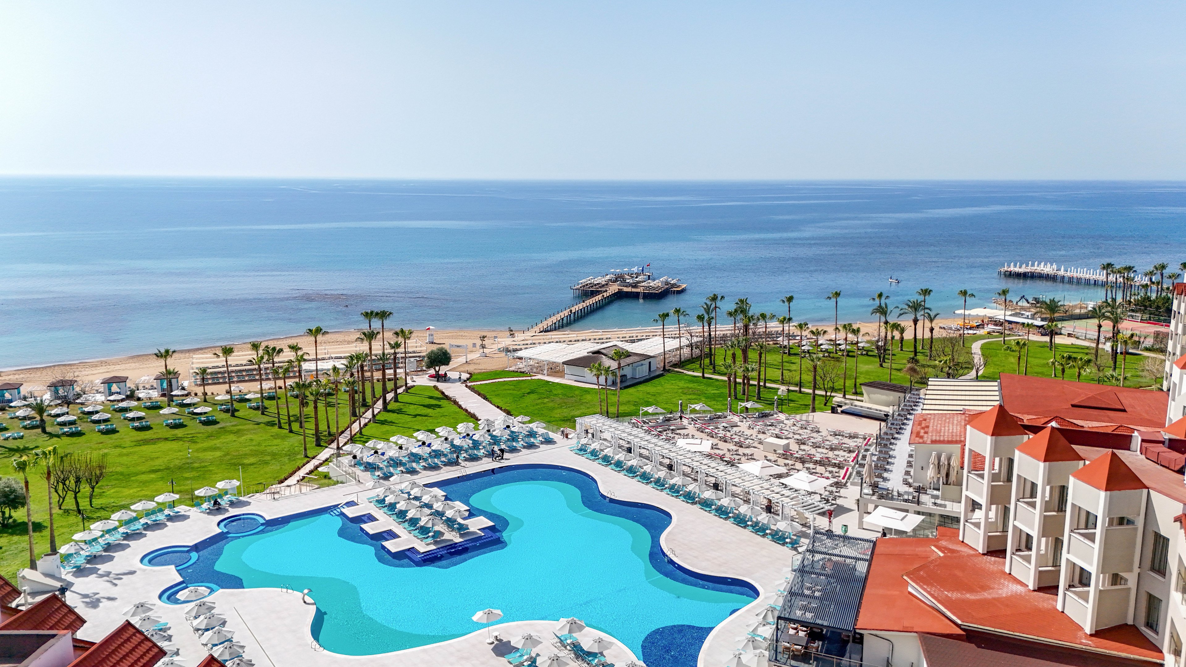 A large resort swimming pool dominates the foreground, surrounded by rows of white sun loungers and umbrellas. Beyond the pool, manicured green lawns with palm trees lead toward a sandy beach. Two long piers extend into the calm blue sea under a clear sky. The scene suggests a sunny, relaxing vacation destination on the coast.