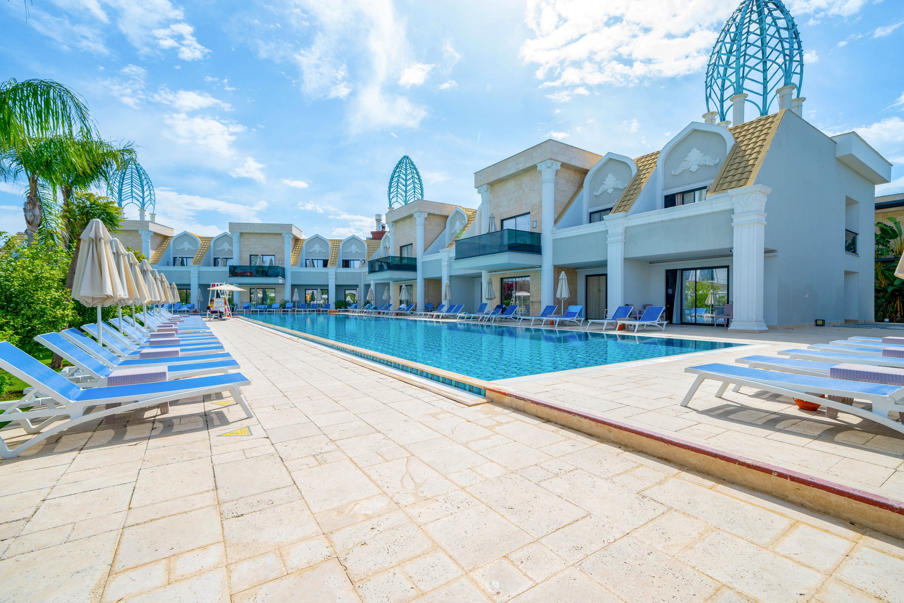 A modern resort features a long outdoor swimming pool lined with white sun loungers and beige umbrellas. Contemporary white buildings with balconies and decorative rooftop structures frame the pool area. Palm trees and greenery add a tropical touch under a bright blue sky with scattered clouds. The scene suggests a relaxing vacation atmosphere at a sunny holiday destination.