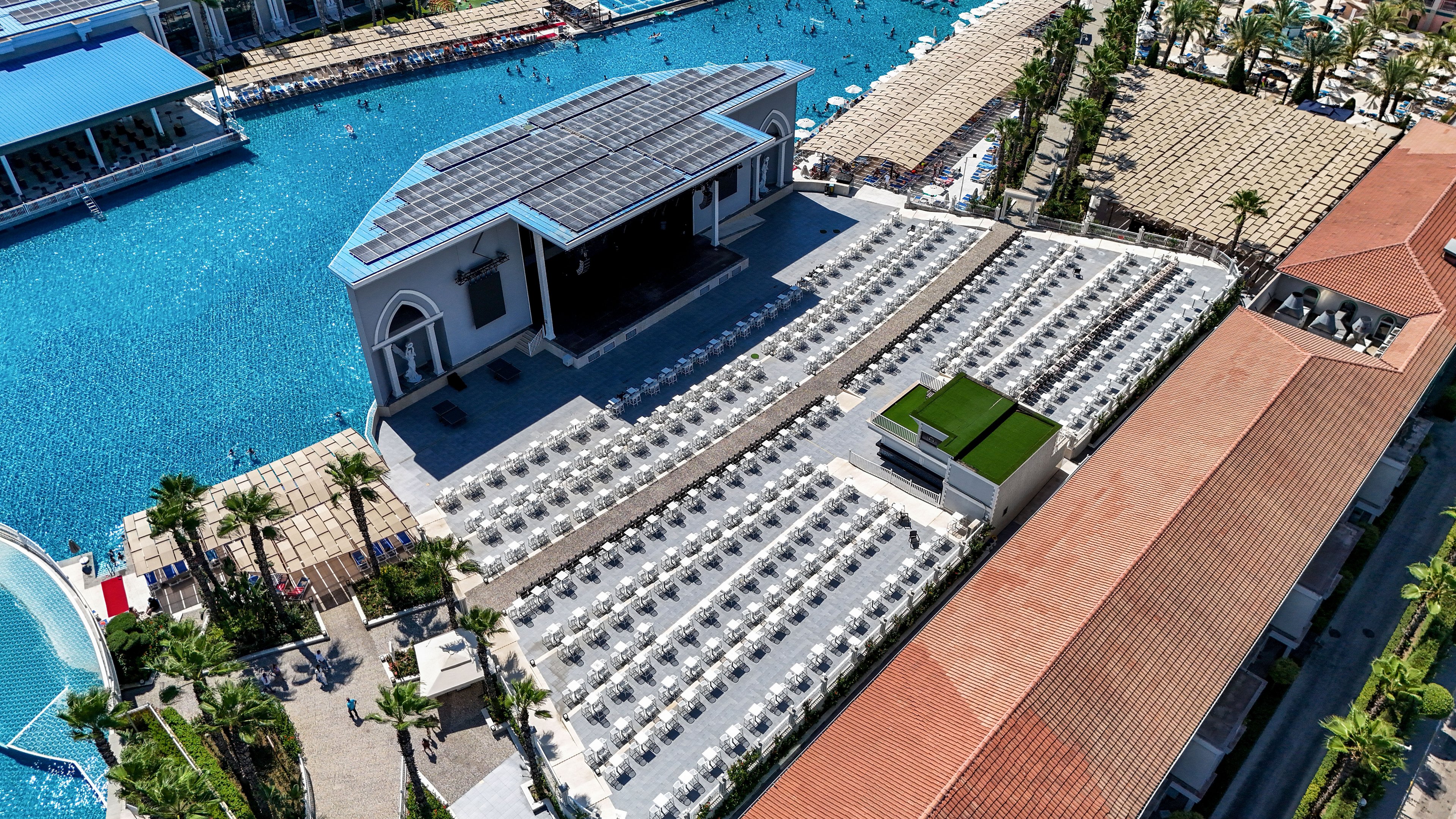 A large outdoor amphitheater with rows of white lounge chairs is shown beside an expansive resort swimming pool. The stage structure features a dark roof and is surrounded by palm trees and landscaped walkways. Guests can be seen enjoying the pool area in the background, adding a lively vacation atmosphere. The scene is captured from above, highlighting the geometric layout and bright, sunny weather.