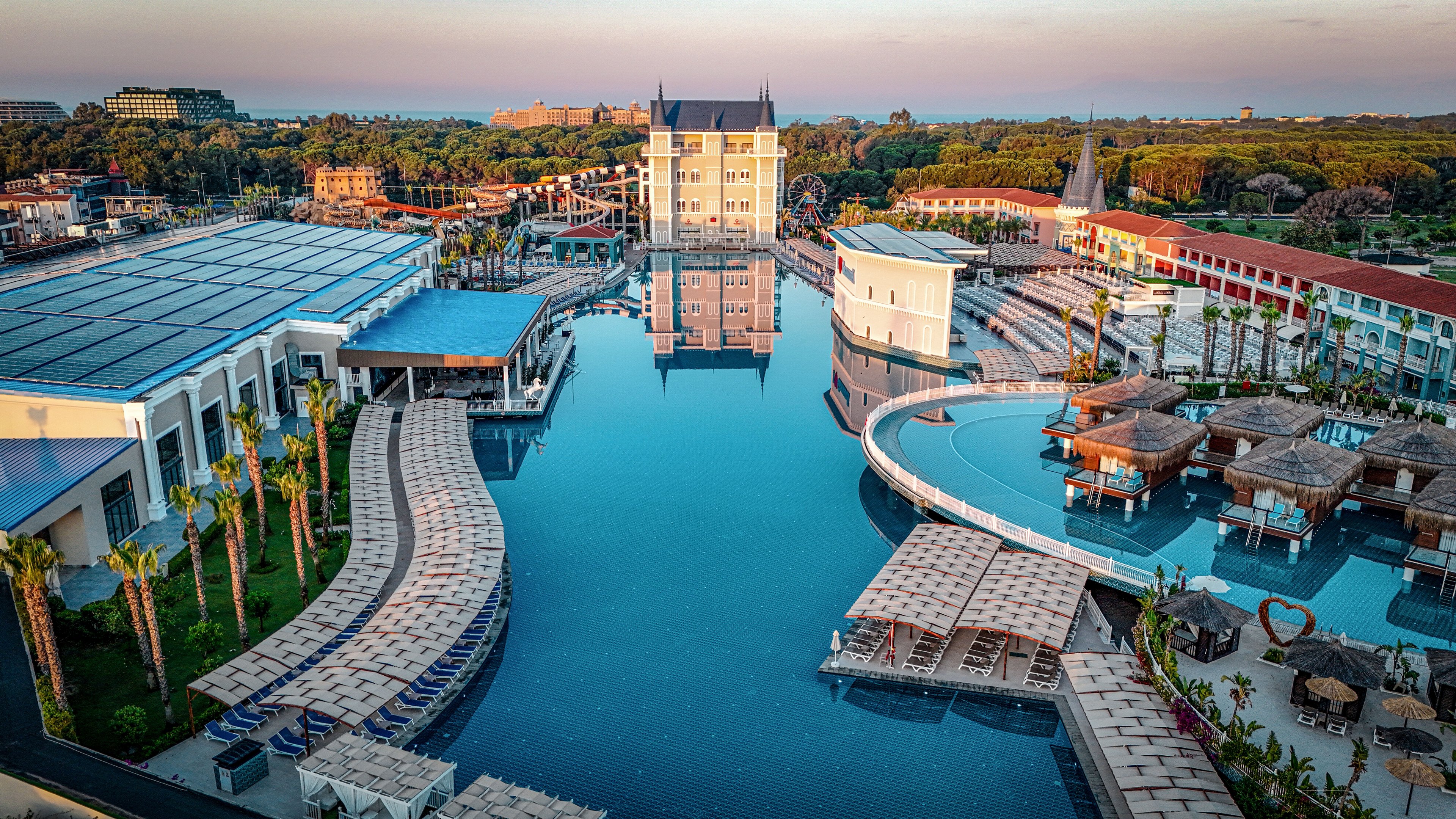 A large luxury resort features an expansive central swimming pool surrounded by sun loungers and cabanas. Modern hotel buildings line both sides of the water, with palm trees adding a tropical atmosphere. The scene is captured at sunset, casting warm light and soft reflections across the calm pool surface. In the distance, greenery and additional structures frame the tranquil vacation setting.