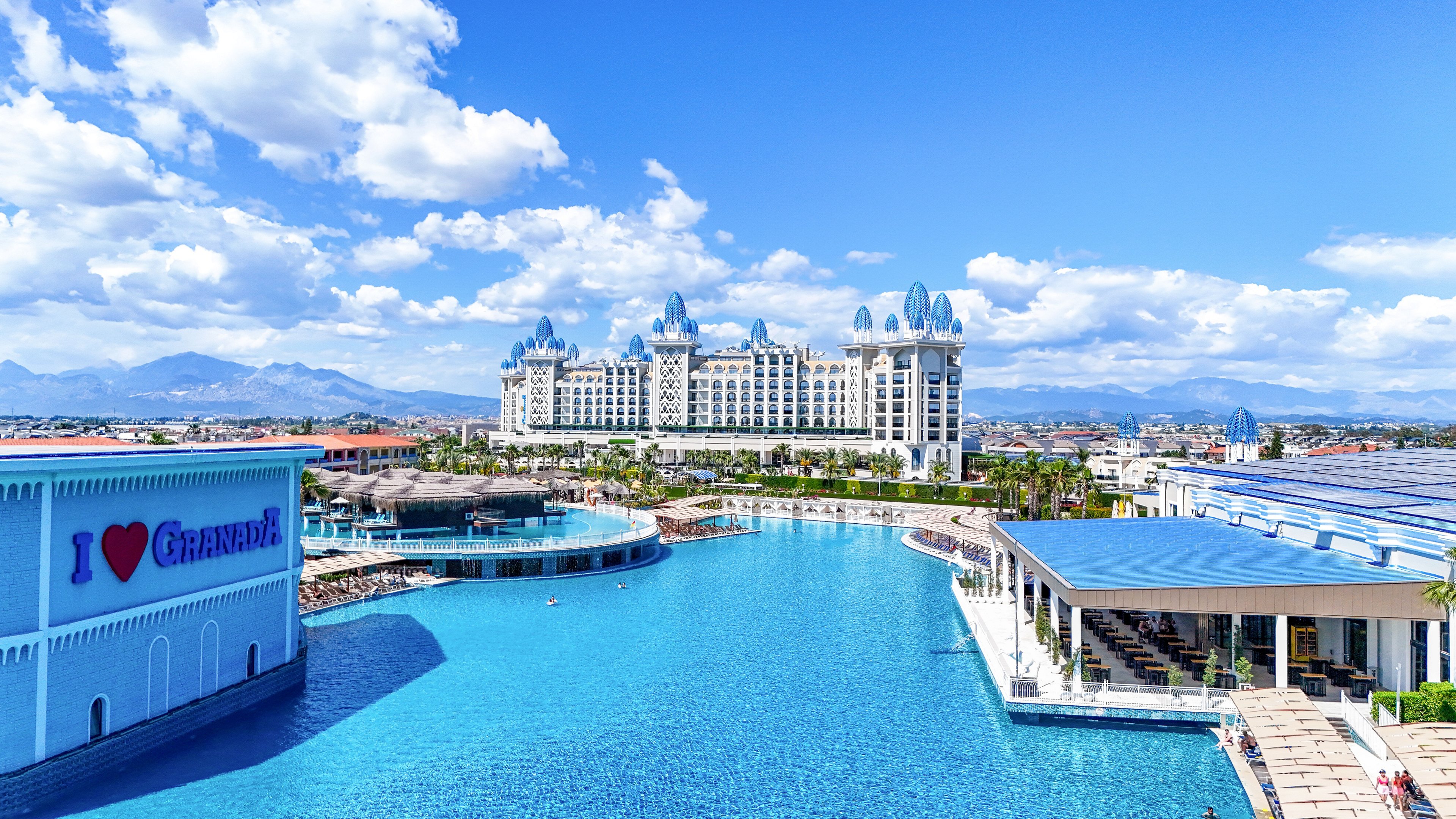 A large, ornate resort hotel with blue-topped towers overlooks an expansive outdoor swimming pool. The scene is set on a sunny day with bright blue sky and scattered clouds, and distant mountains visible on the horizon. A building by the pool displays the text "I ♥ GRAND" in bold colors, adding a playful touch to the leisure setting. Palm trees, poolside walkways, and sunlit terraces emphasize a luxurious vacation atmosphere.