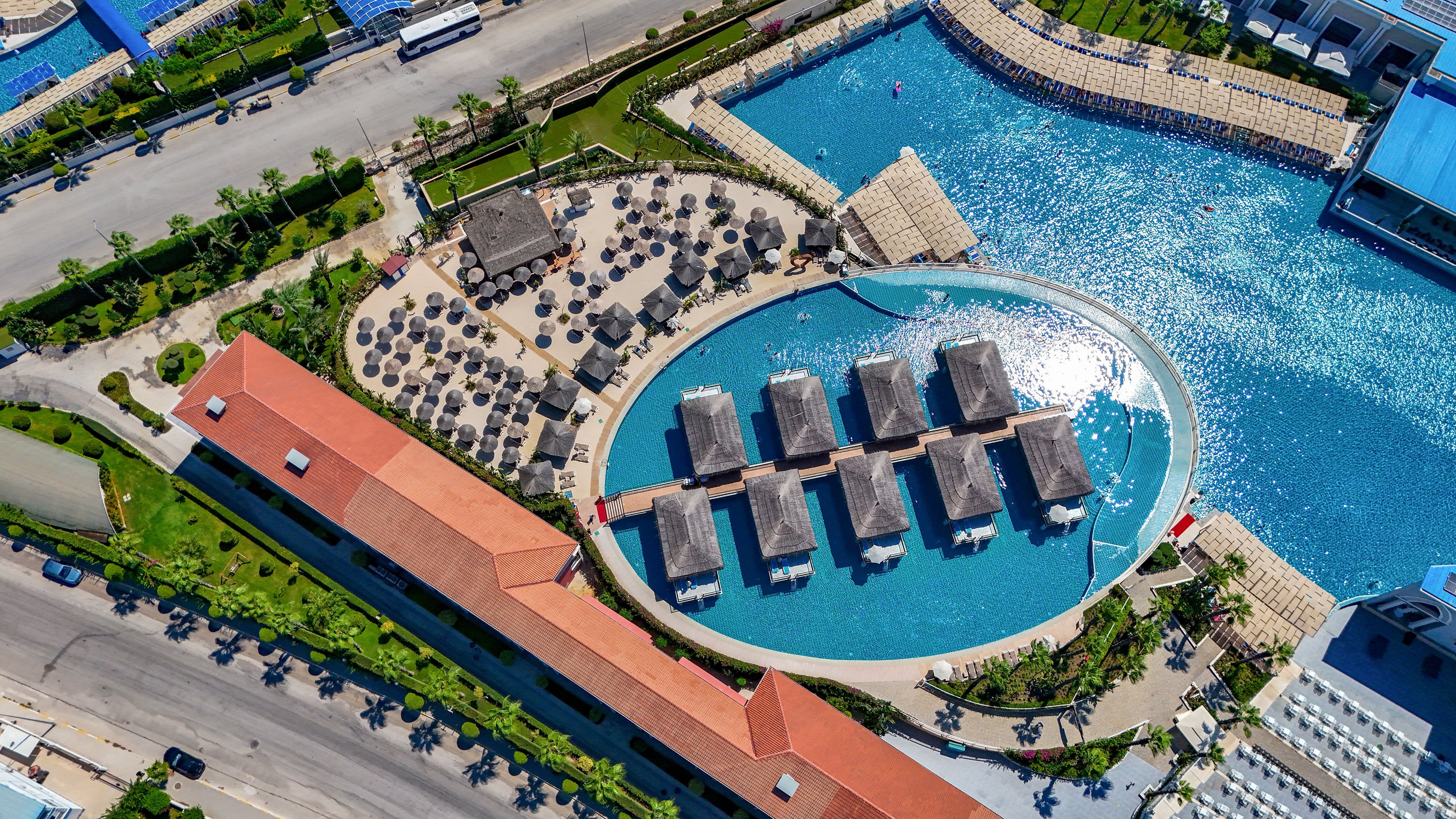 A large resort pool complex is captured from above, showing an oval lagoon with overwater cabanas and surrounding sun loungers. The pool sits beside a bright blue waterfront, separated by a curved stone edge and landscaped walkways. Rows of umbrellas and chairs create a structured pattern, while nearby buildings and a road frame the leisure area. The scene conveys a sunny, tropical vacation atmosphere with clear turquoise water and organized outdoor spaces.