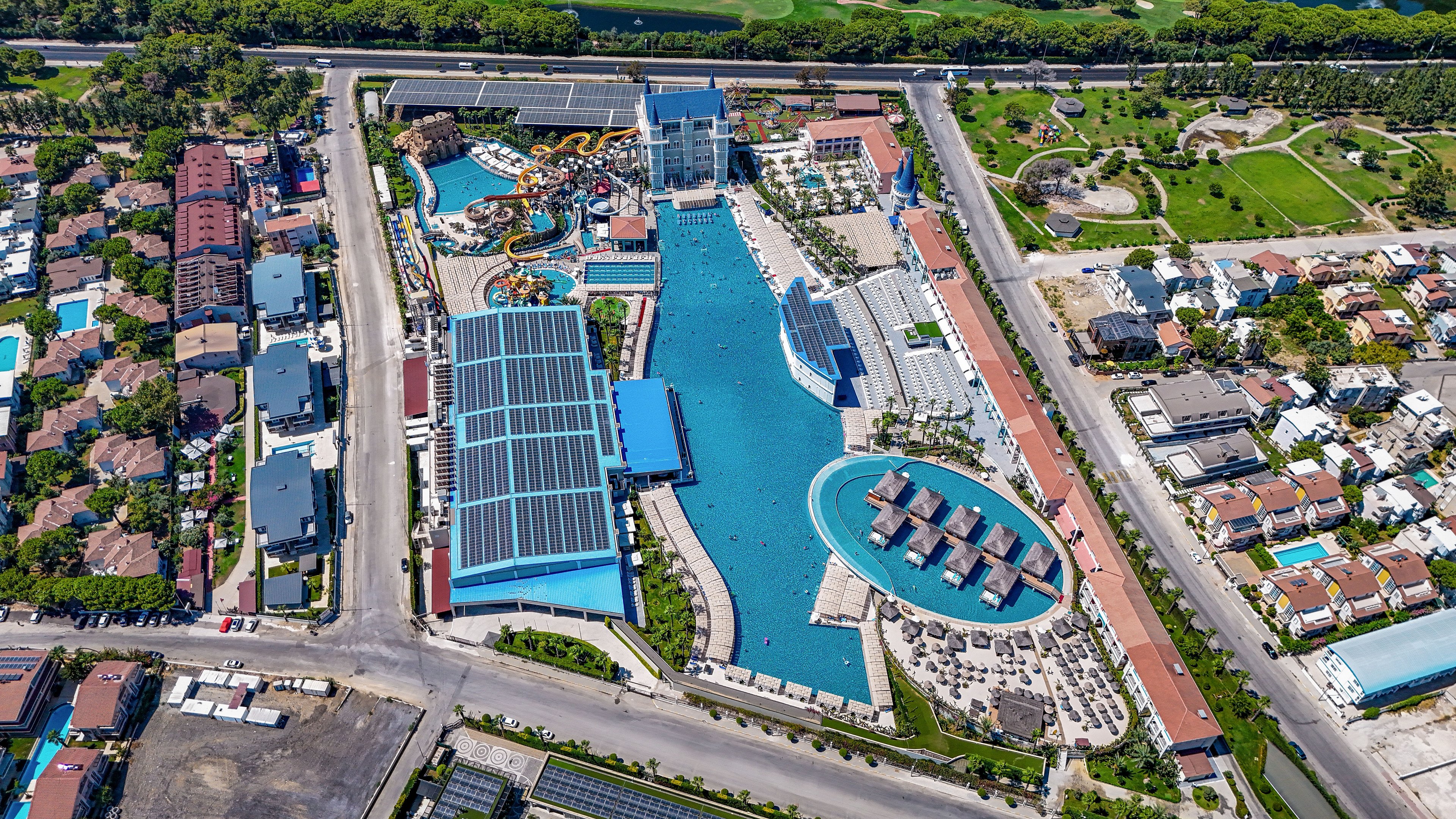 A large resort complex with an expansive central pool and waterpark is captured from above. Multiple waterslides, play structures, and shaded lounge areas surround the turquoise swimming pools. Solar panel-covered buildings line one side, while residential houses and green park spaces border the property. The scene is bright and sunny, highlighting the organized layout of pools, pathways, and recreational zones.