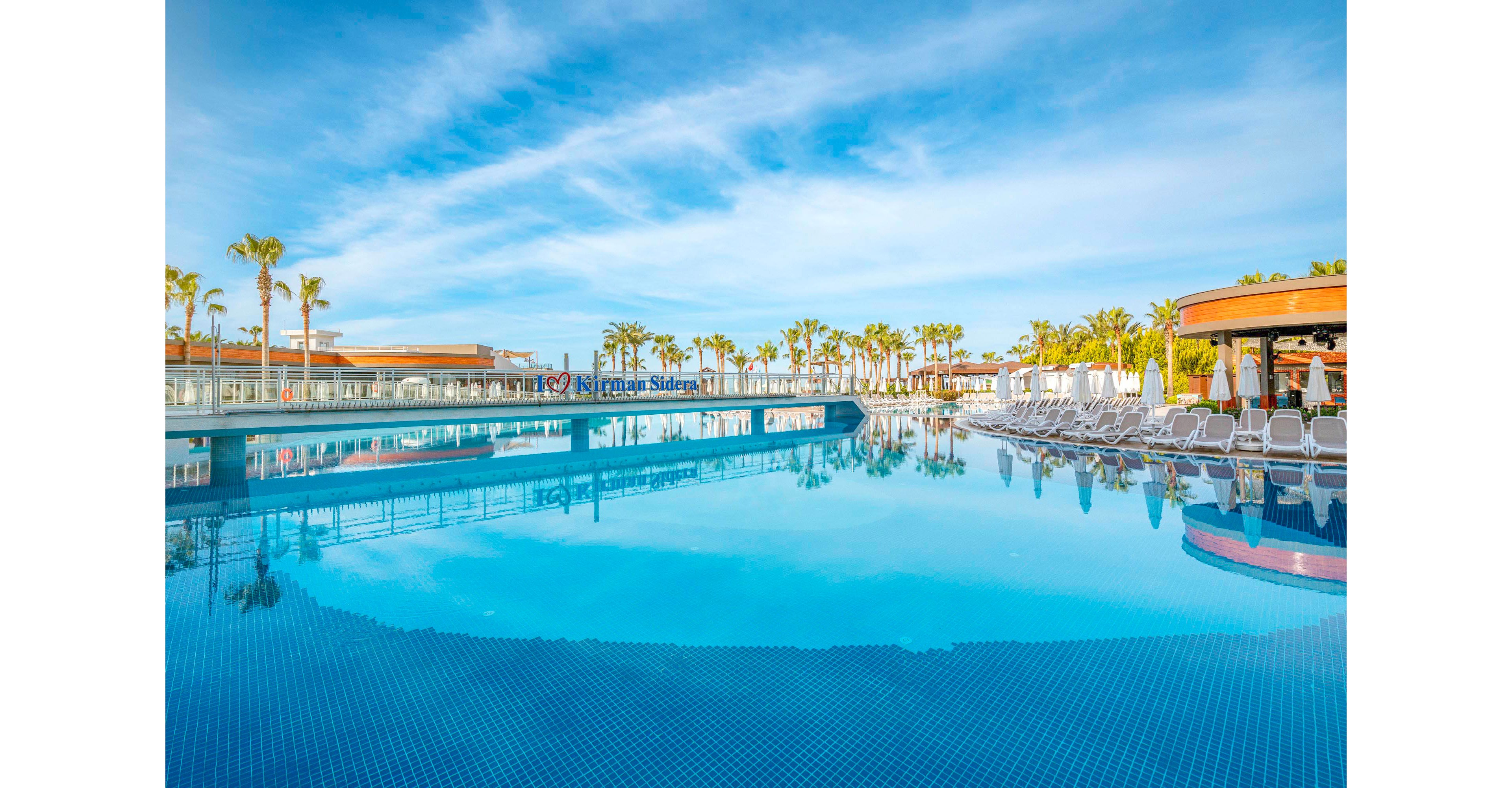 A large, tranquil swimming pool is the main focus, surrounded by rows of white lounge chairs and tall palm trees. The setting appears to be a modern resort under a bright blue sky with scattered clouds. The pool reflects the surrounding architecture and greenery, creating a serene and inviting atmosphere. The visible text 'Riu Palace' is displayed on a sign in the background.